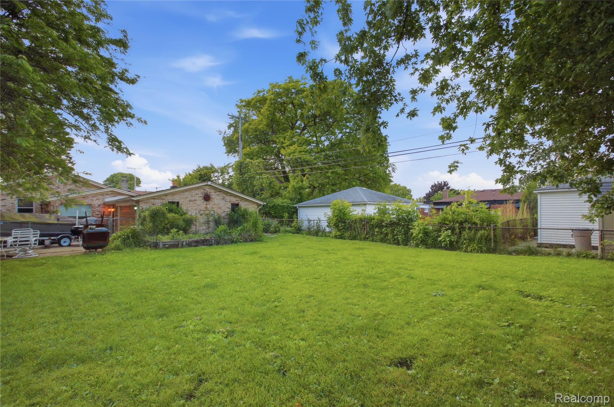 Grassy backyard behind a brick house, with garden beds, mature trees, and a chain-link fence.