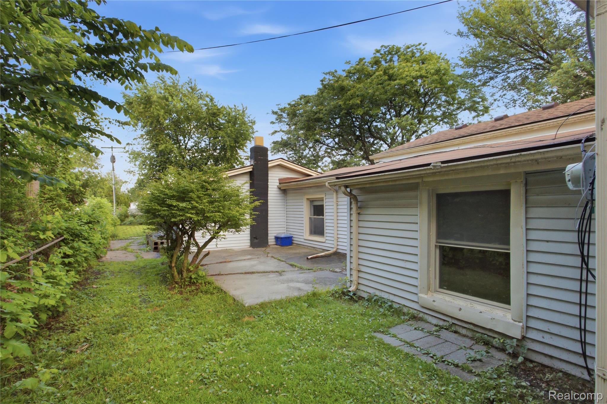 House side yard with a paved patio, grassy lawn, mature trees, and a dark chimney.