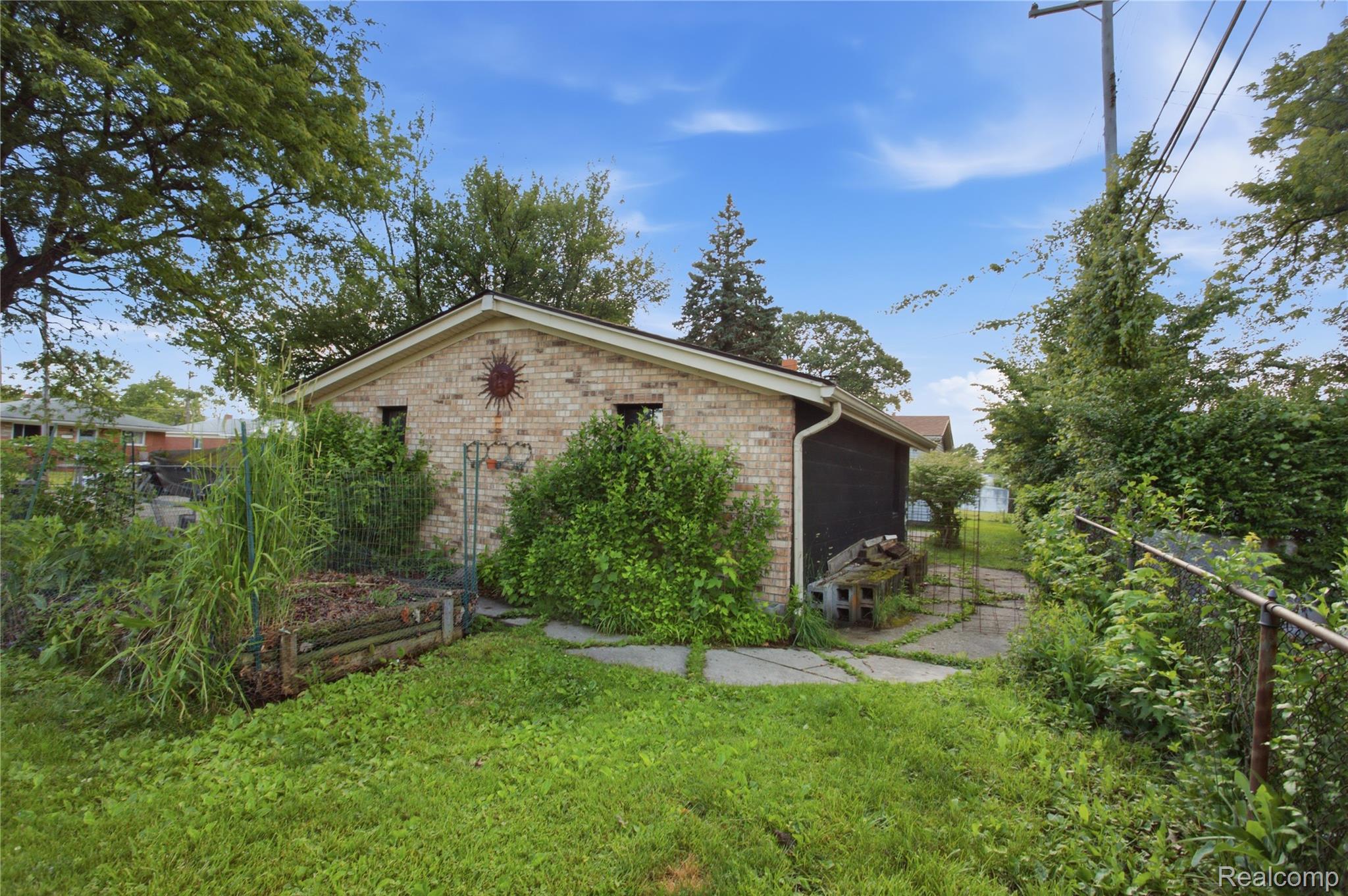 Brick garage with a dark door, overgrown garden beds, and a paved walkway through the yard.