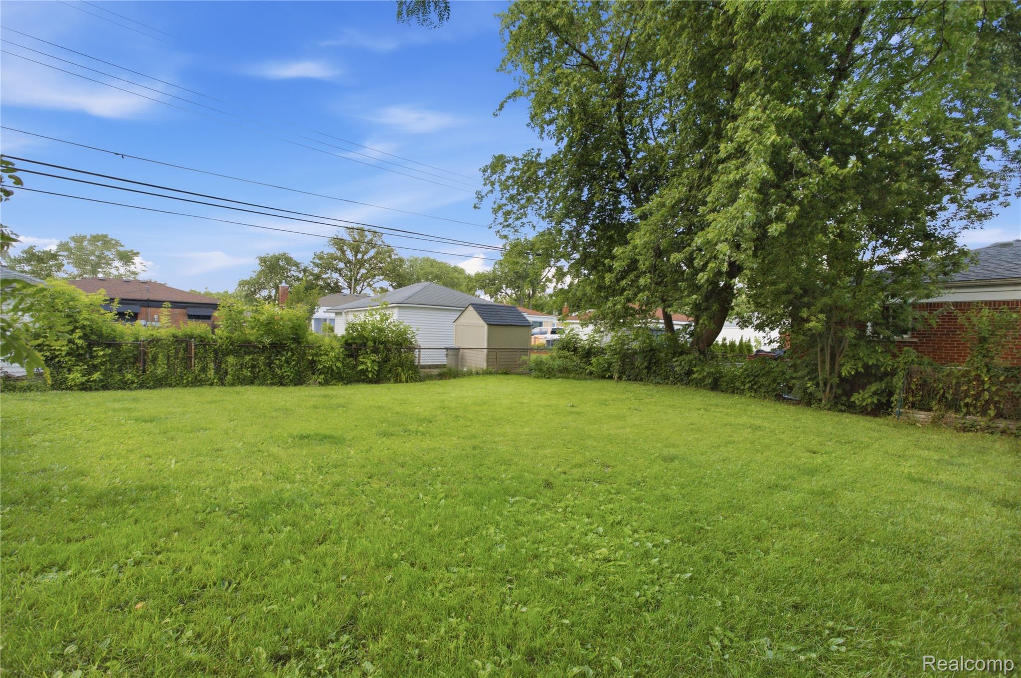 Backyard lawn bordered by mature trees, shrubs, and a fence, with neighboring homes beyond.