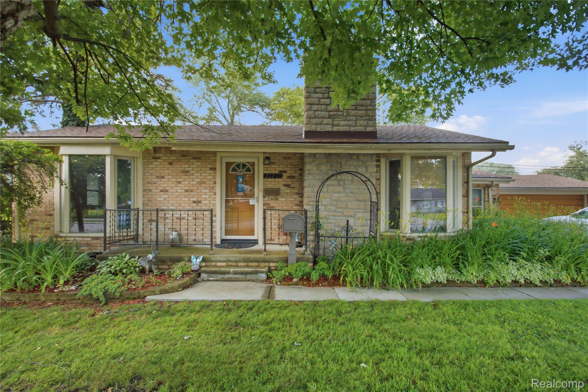 Brick house with a stone chimney, covered entry, and mature trees shading the landscaped front yard.