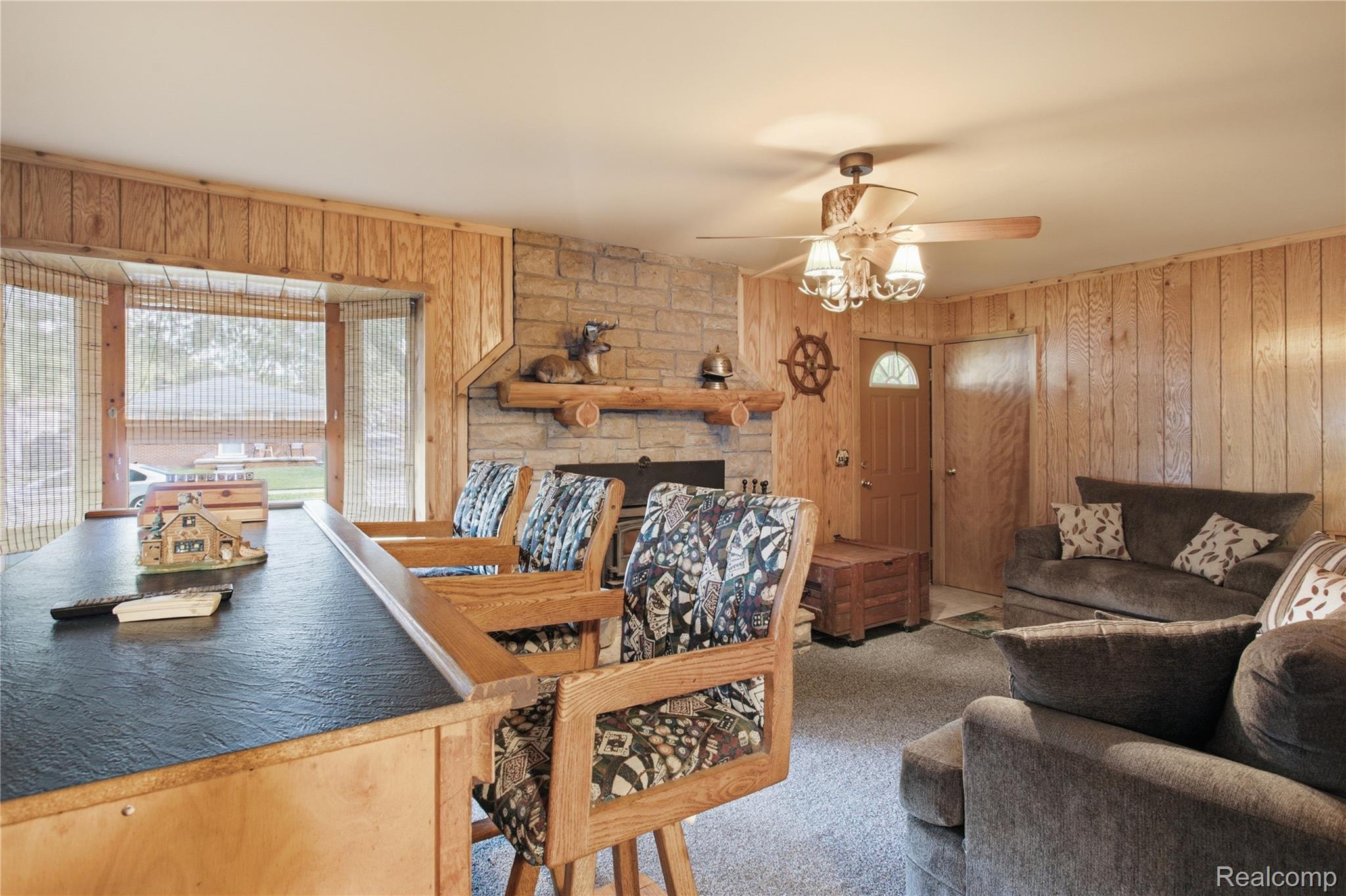 Living and dining room with a stone fireplace, wood-paneled walls, and patterned dining chairs.
