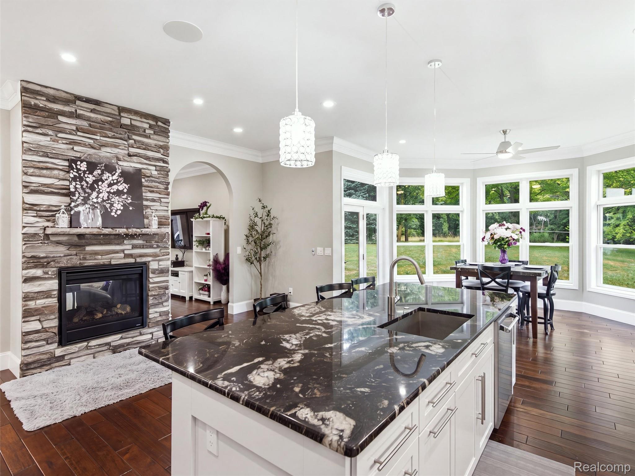 Open kitchen with a large dark stone island, stone fireplace, and dining area beside wide windows.