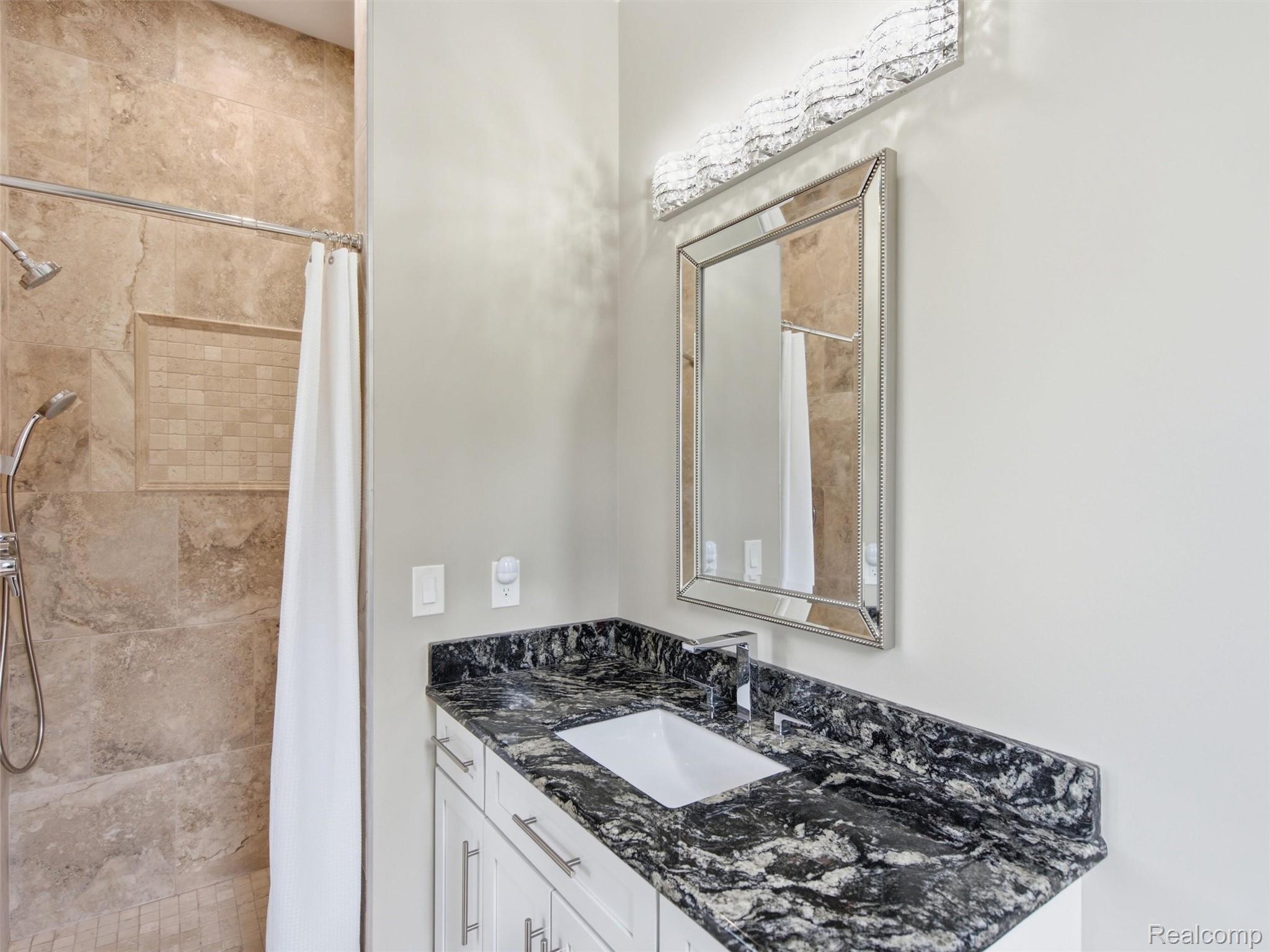 Bathroom with a beige tiled shower and white vanity topped with dark veined granite.