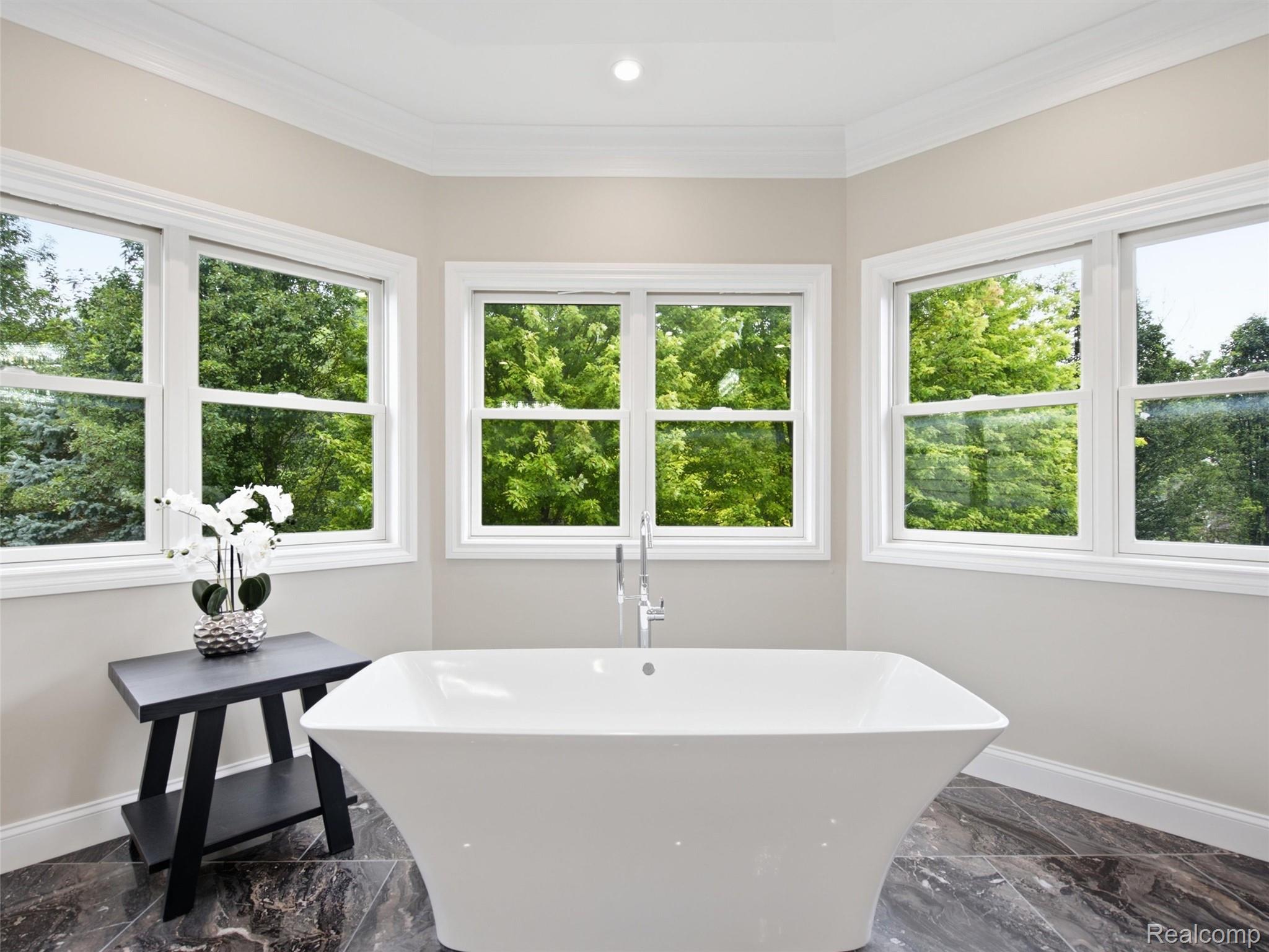Bathroom with a freestanding soaking tub beneath large windows overlooking green trees.