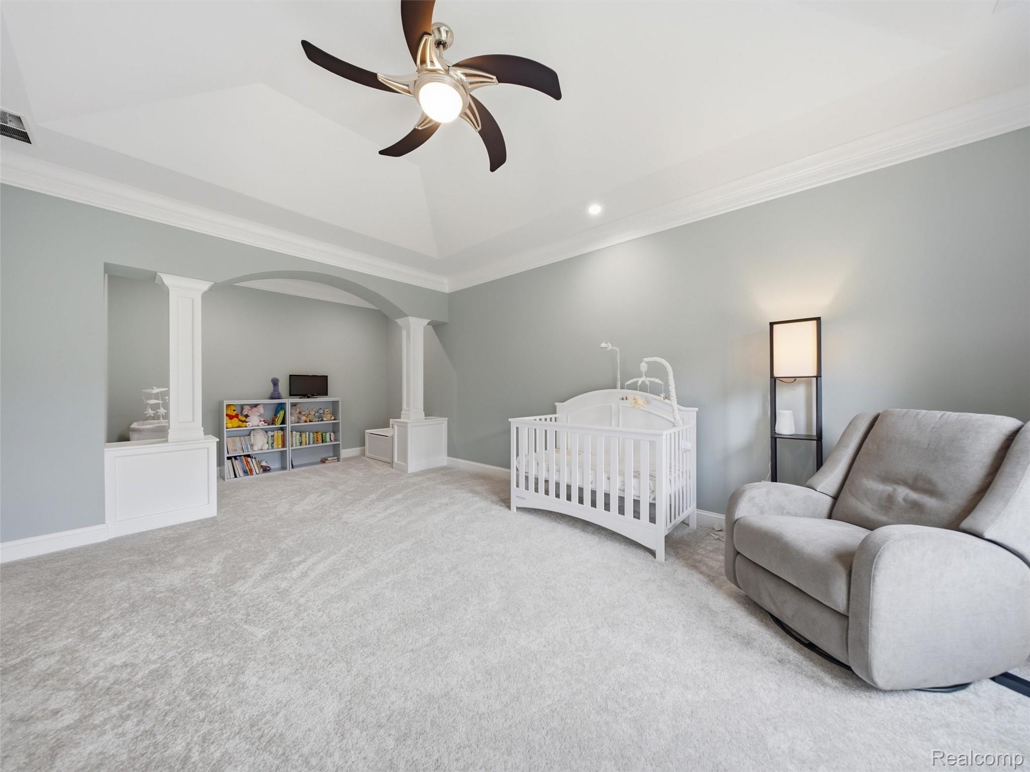 Nursery with a white crib, gray glider, and toy shelves beneath a tray ceiling.