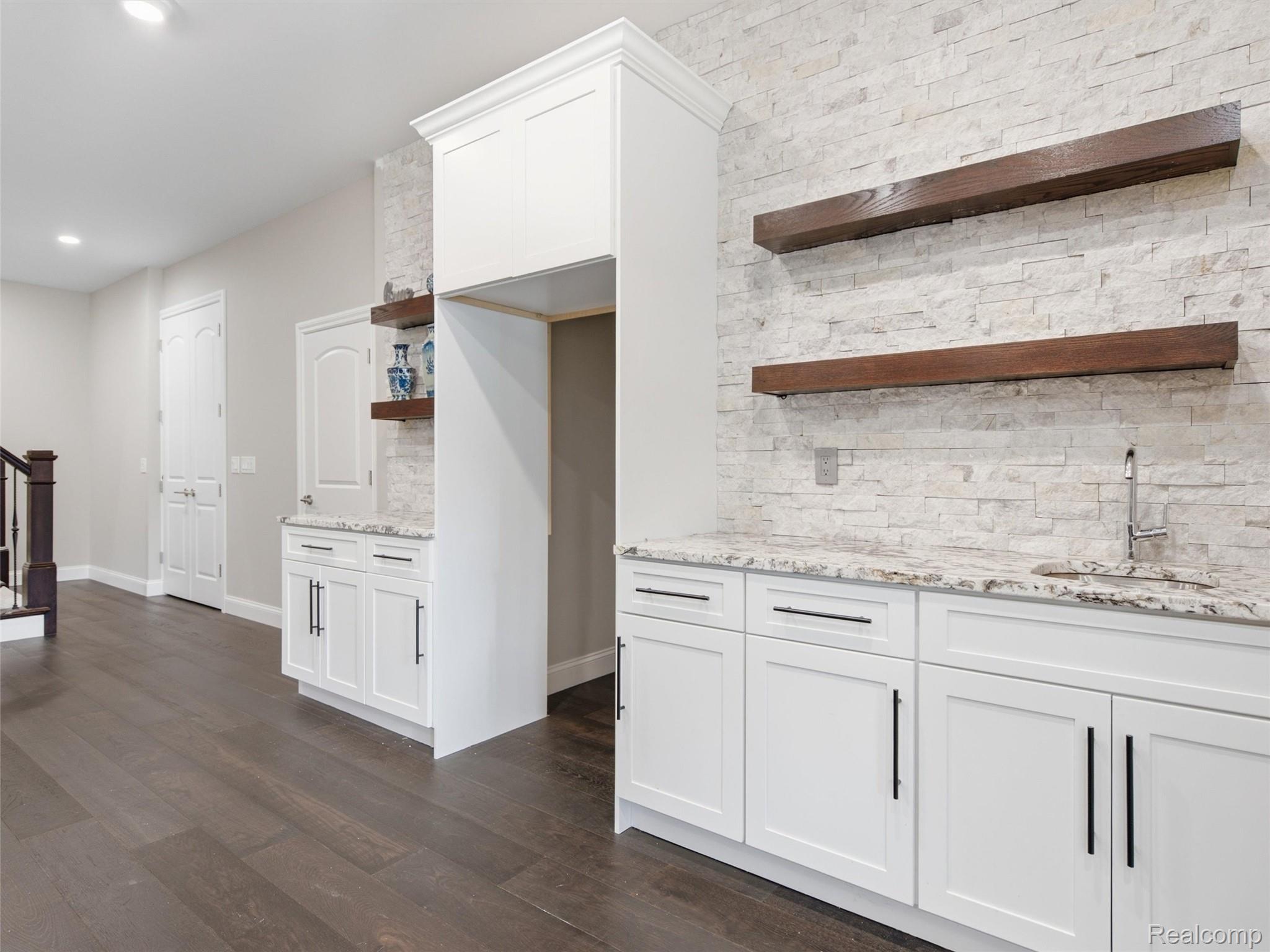 Kitchen cabinetry with a stone backsplash, dark floating shelves, granite counters, and a stainless faucet.