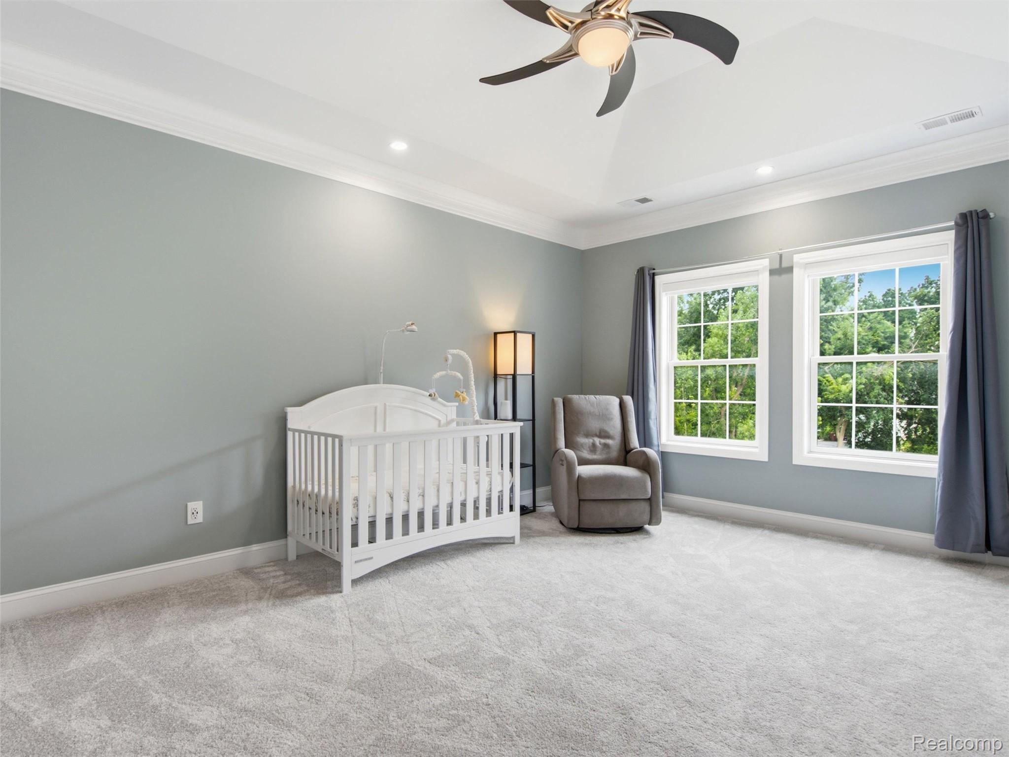 Nursery with a white crib, gray rocking chair, and two windows overlooking leafy trees.