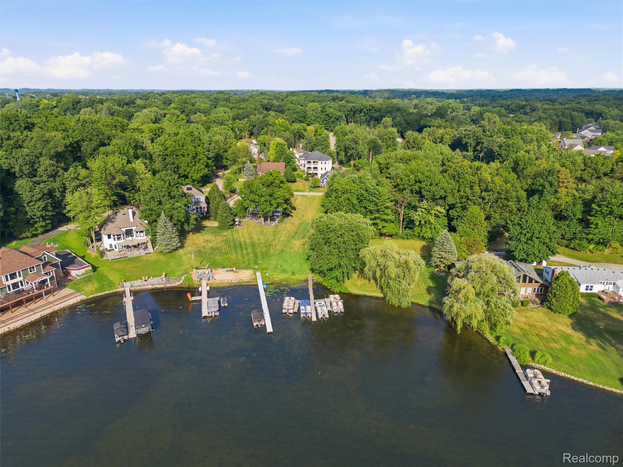 Aerial view of homes along a wooded lake shoreline, with green lawns and several docks.