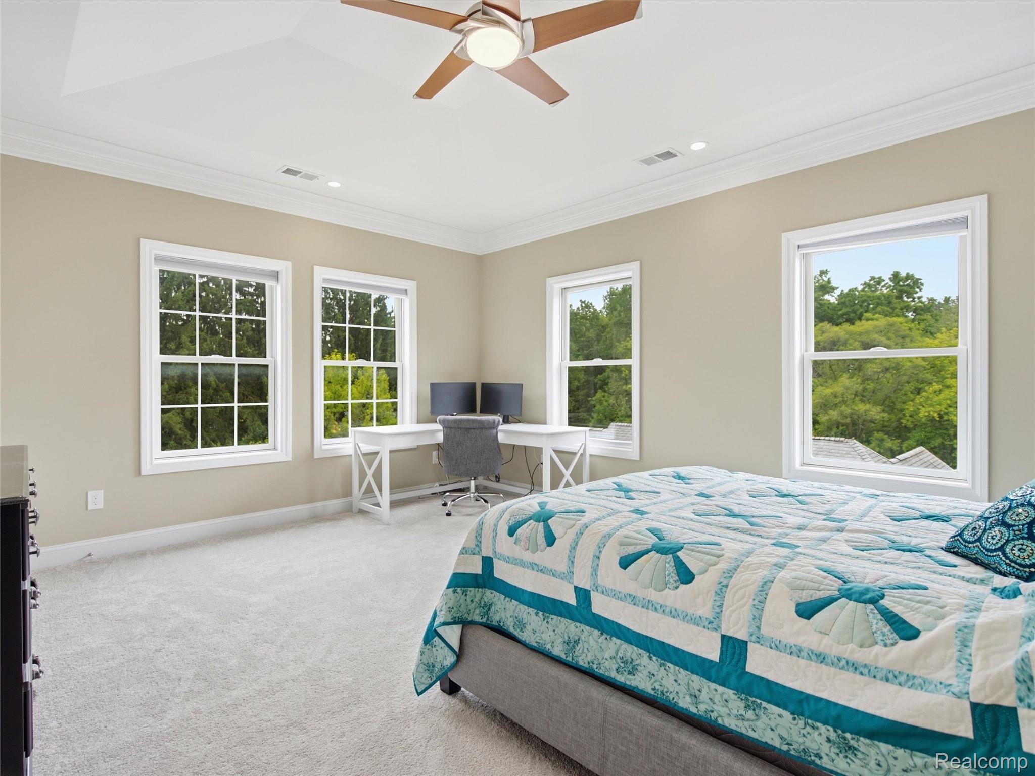 Bedroom with a blue-and-white quilted bed, corner desk, ceiling fan, and windows overlooking trees.