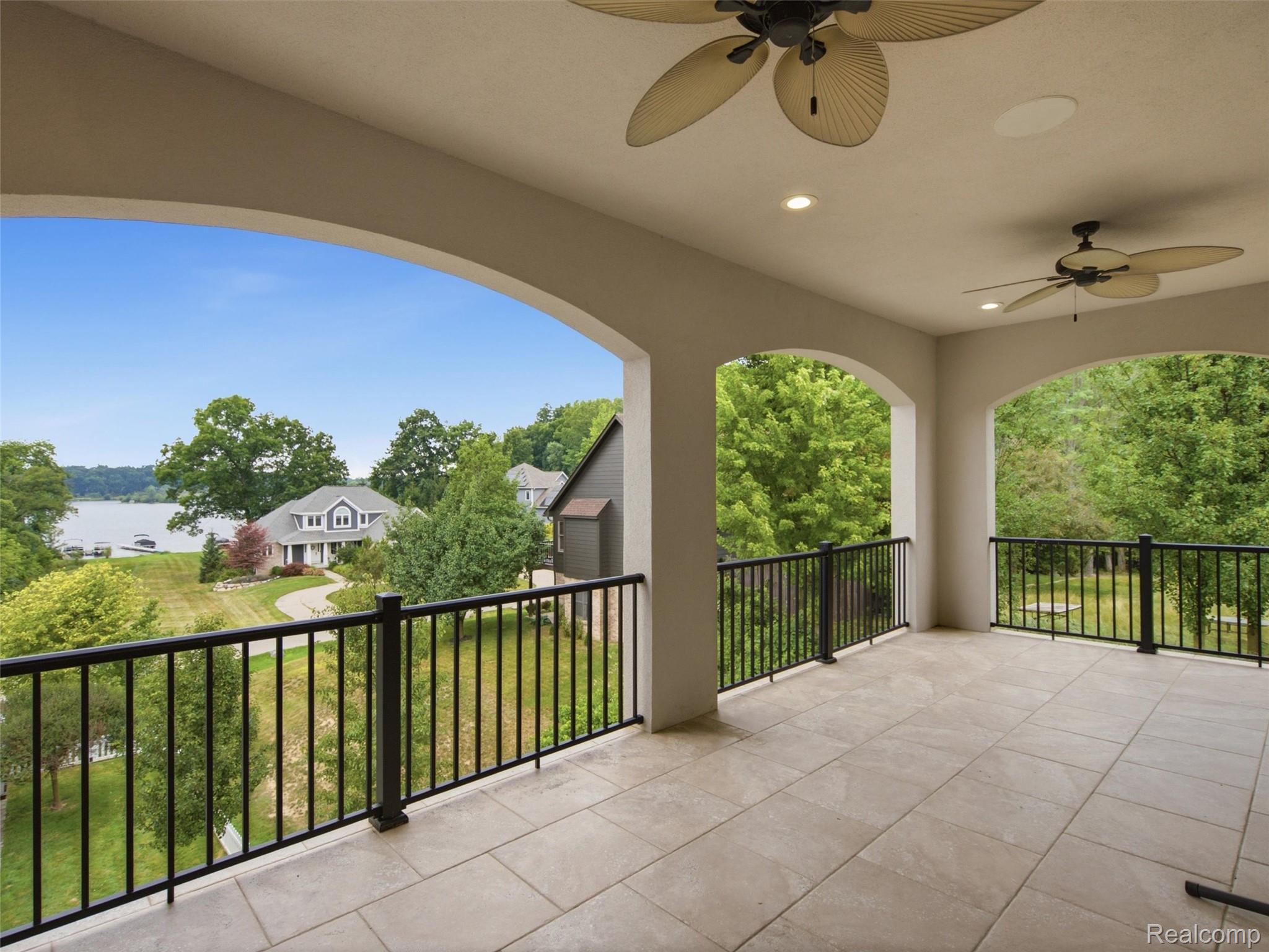 Covered balcony with arched openings and ceiling fans overlooking a lake and neighboring homes.