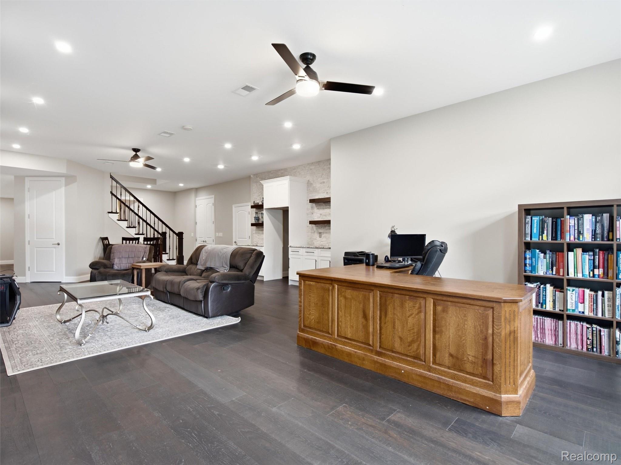 Open-plan living room and office with gray recliners, a large wooden desk, and dark flooring.