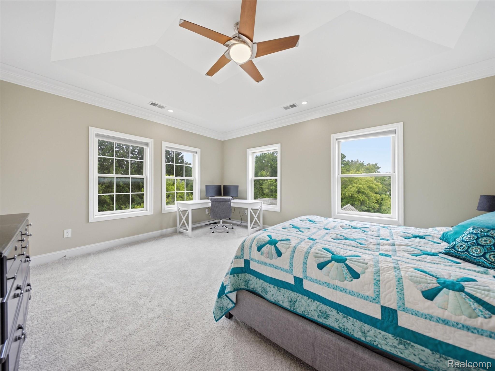 Bedroom with a teal patterned quilt, corner desk, and windows overlooking trees.