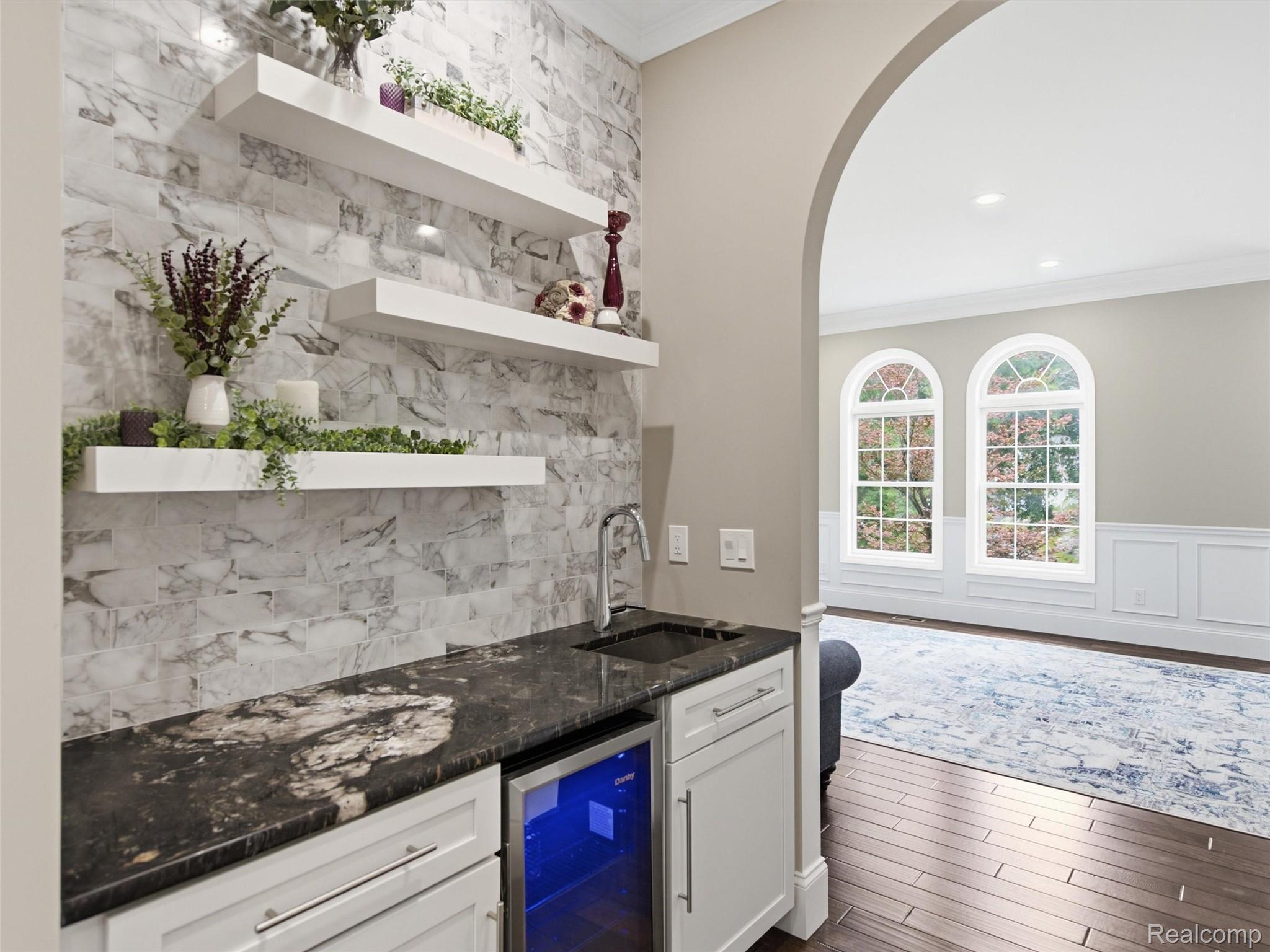 Built-in wet bar with marble-look backsplash, floating shelves, sink, and beverage refrigerator beside an arched sitting room.