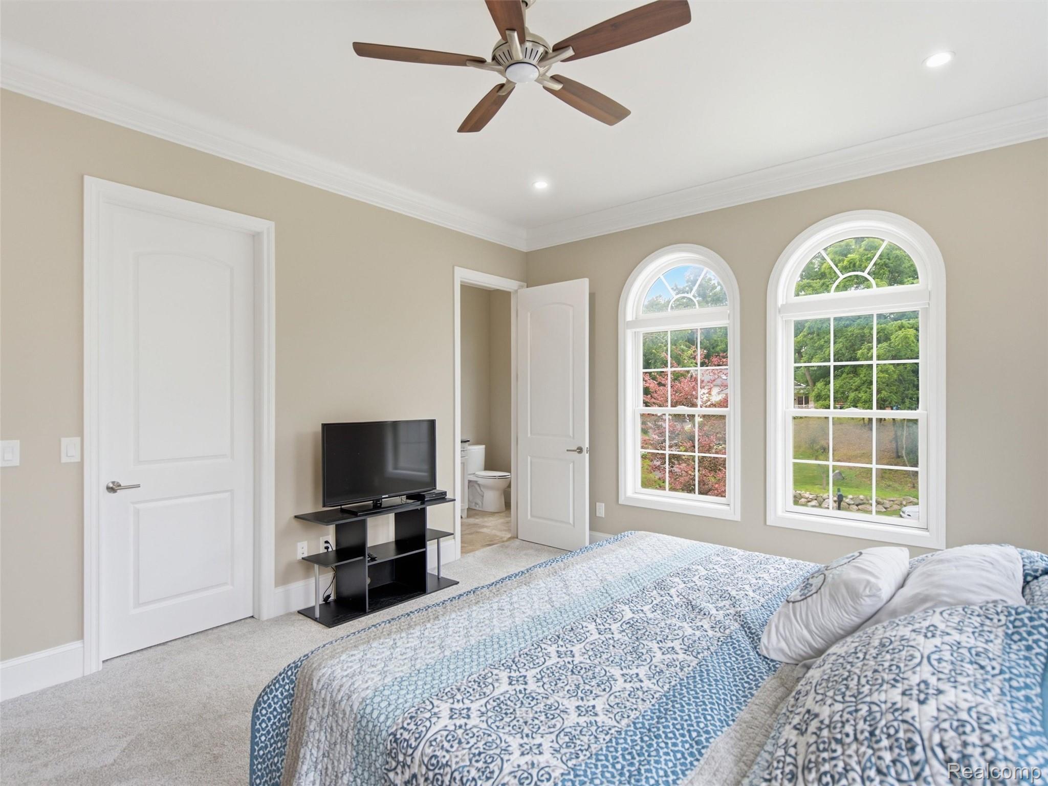 Bedroom with a patterned bed, ceiling fan, and two arched windows overlooking greenery.