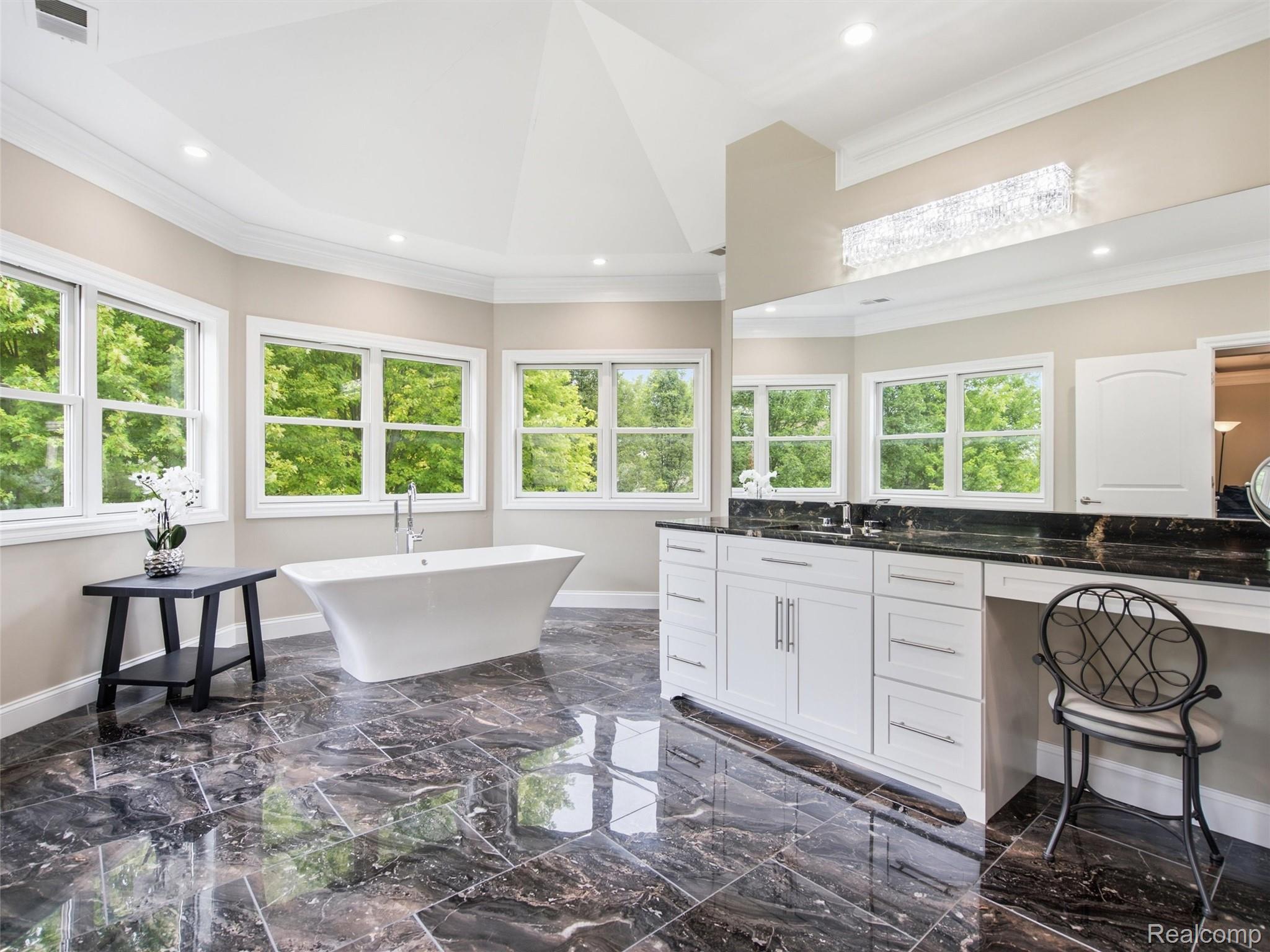 Bright bathroom with a freestanding soaking tub, tree-view windows, white vanity, and glossy dark tile floor.