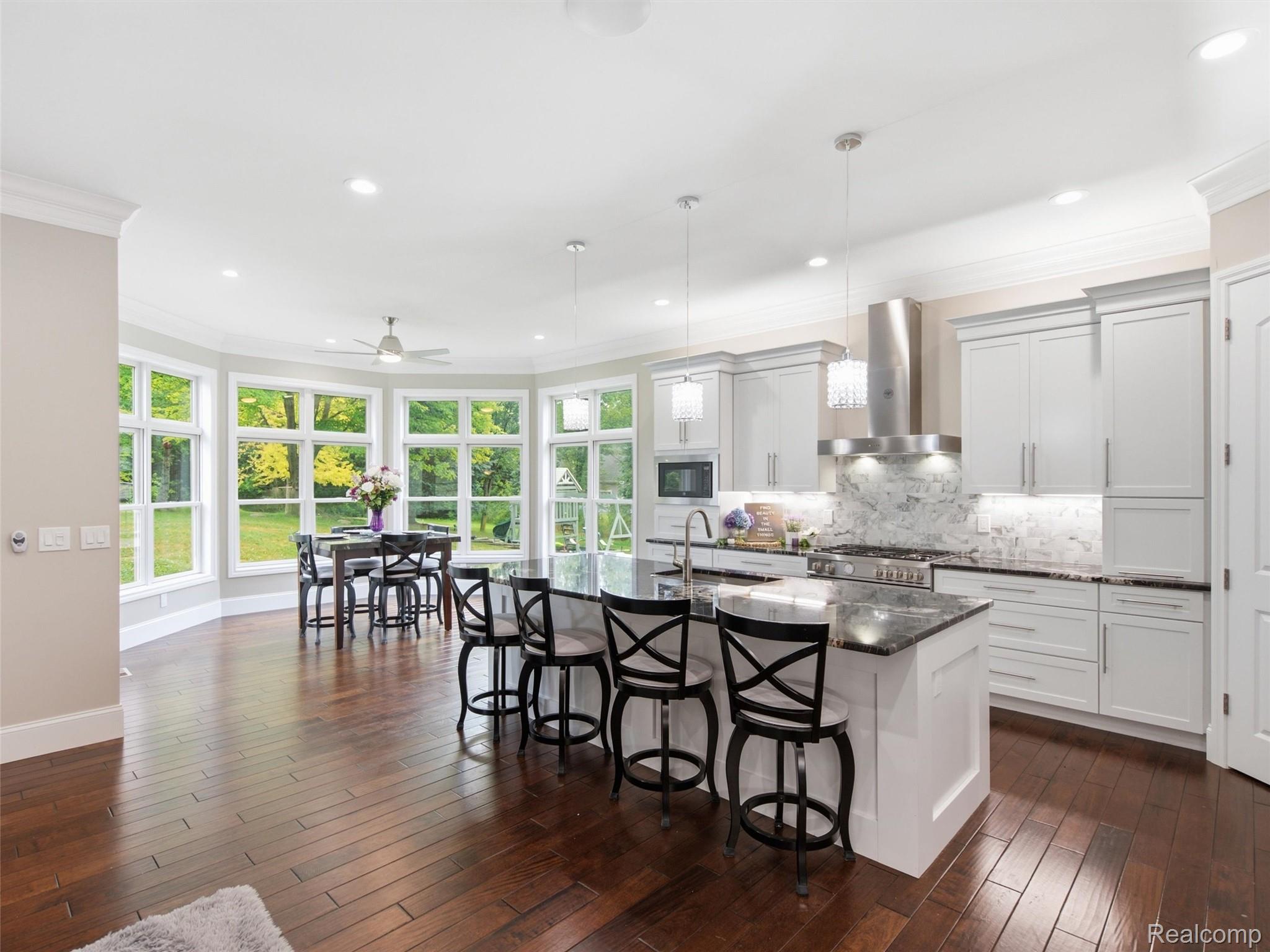 Kitchen with a large stone island, black bar stools, white cabinets, and windows overlooking a green yard.