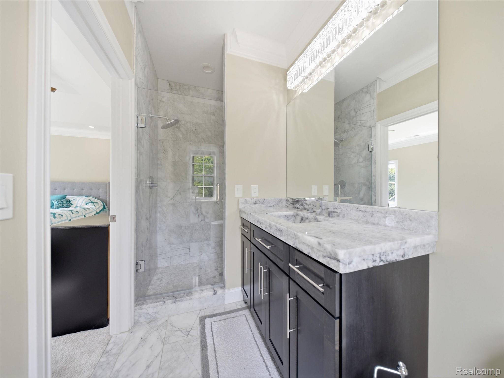 Bathroom with a marble-tiled glass shower and dark vanity, with a bedroom visible through the doorway.