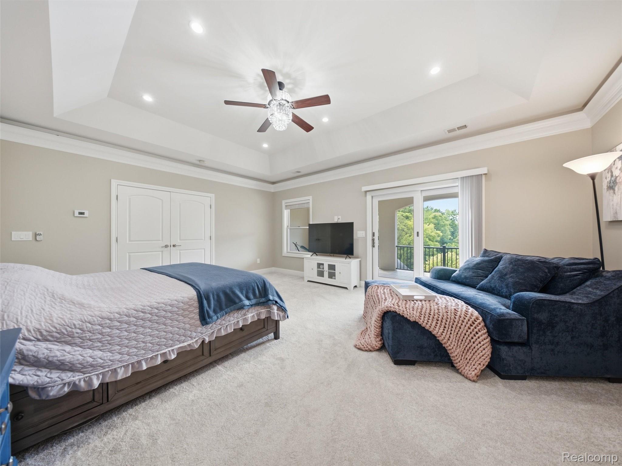 Furnished bedroom with a sitting area, tray ceiling, and sliding door to a tree-lined balcony.