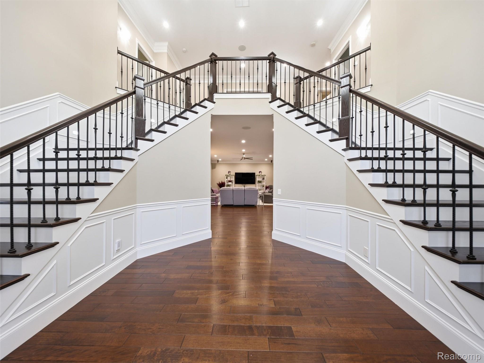 Entry hall with twin staircases, dark railings, white paneled walls, and wood flooring.