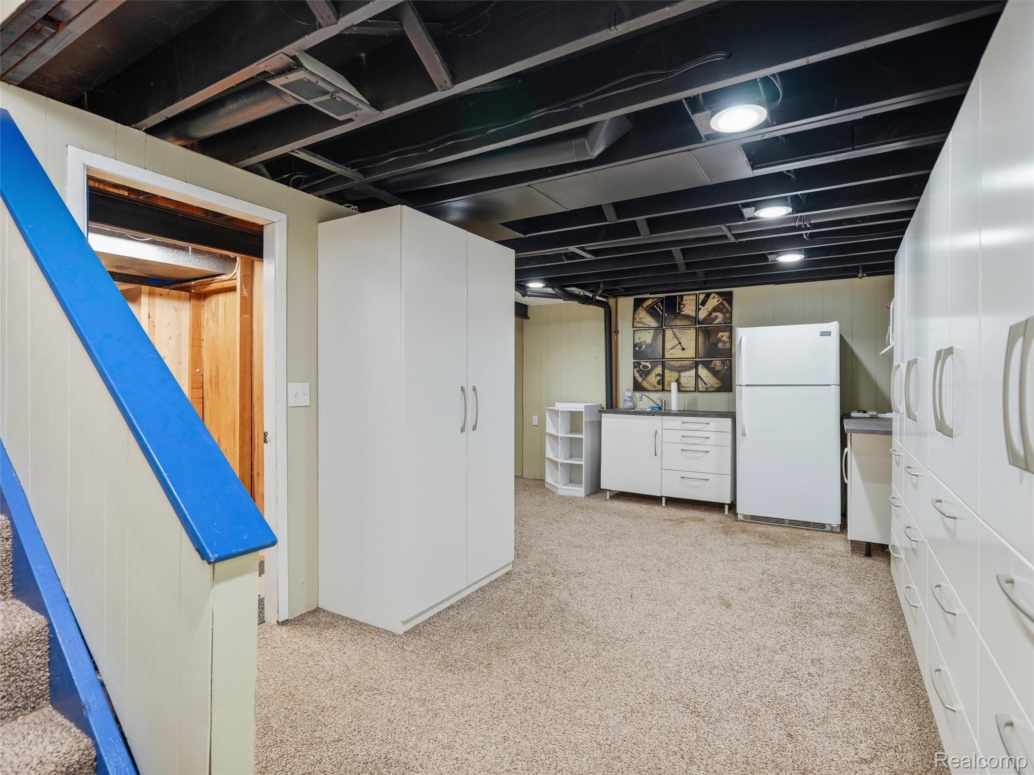 Basement utility area with white storage cabinets, a refrigerator, and blue stair railings.