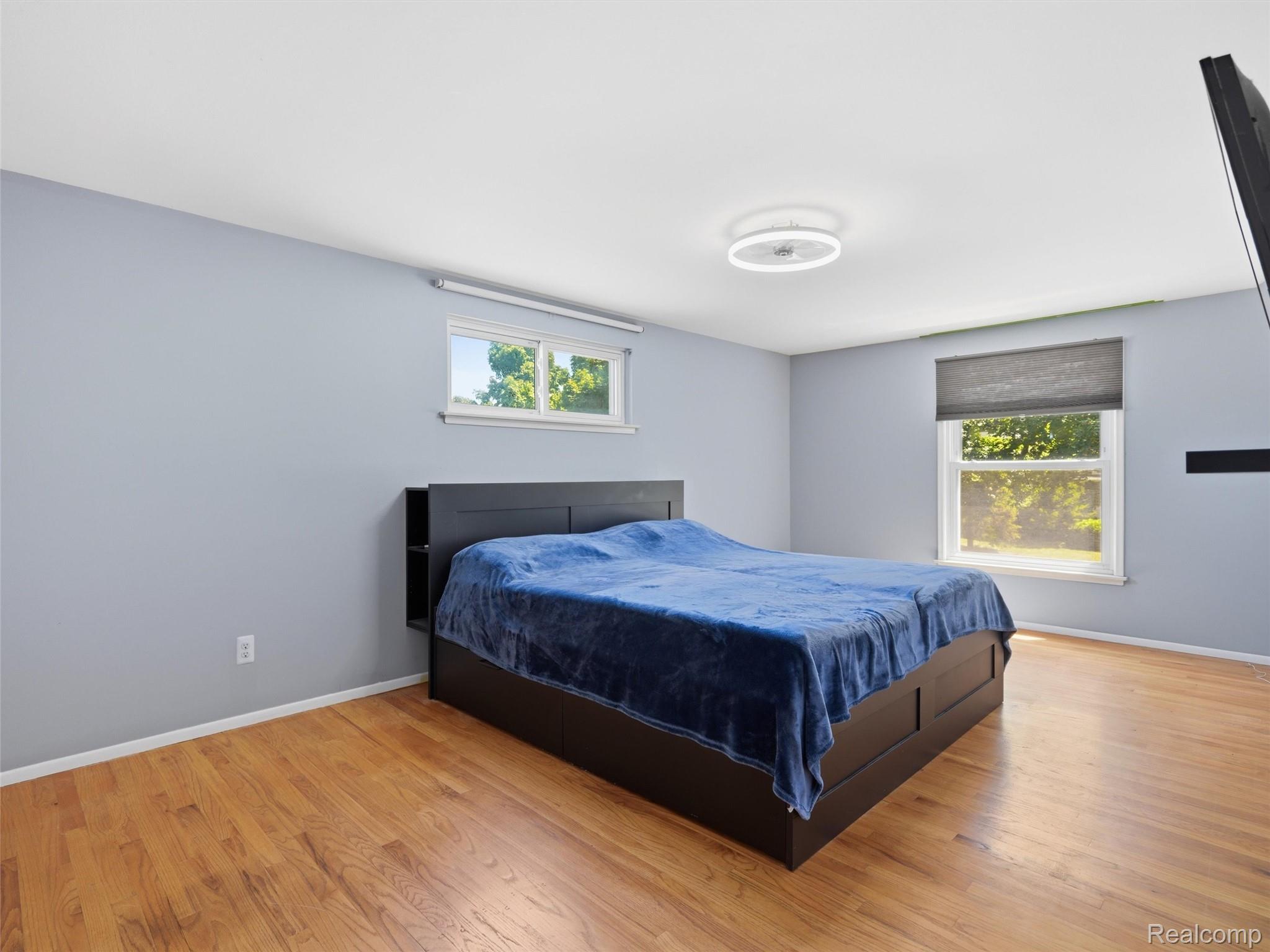 Bedroom with a dark bed and blue blanket, hardwood floors, and two windows.