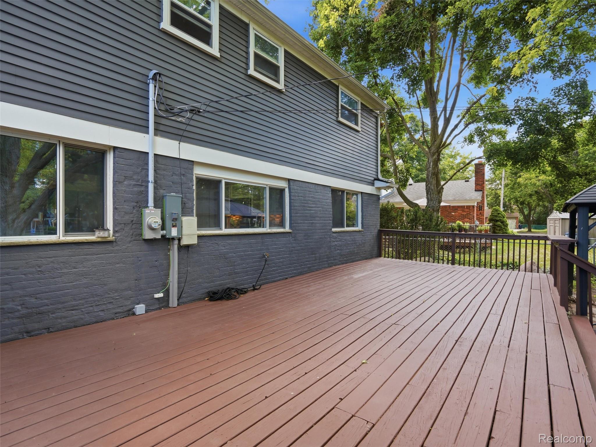 Wide brown deck beside a gray-sided house, with dark railings and mature trees beyond.