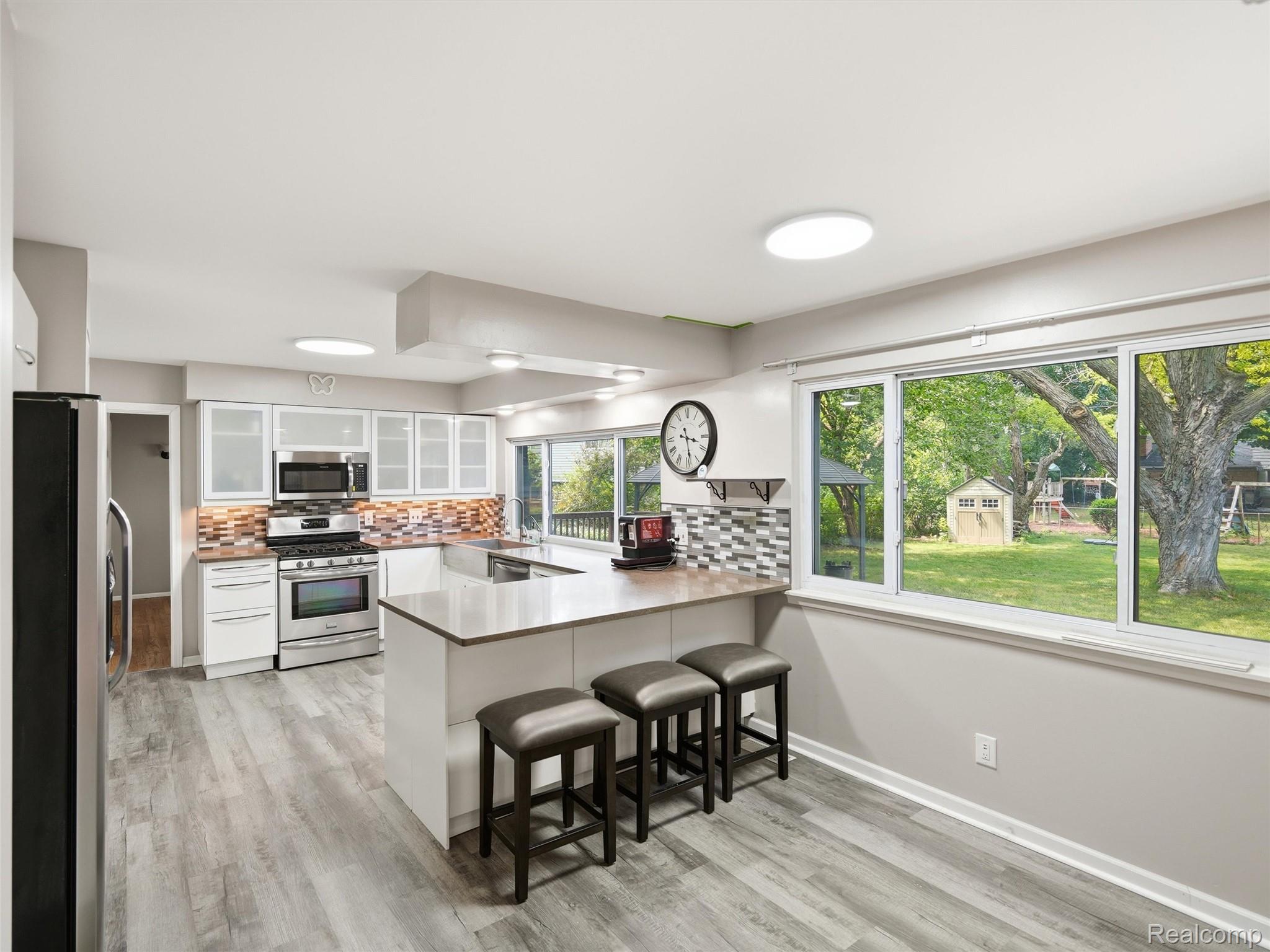 Kitchen with a large island and three stools, opening to a leafy backyard through wide windows.