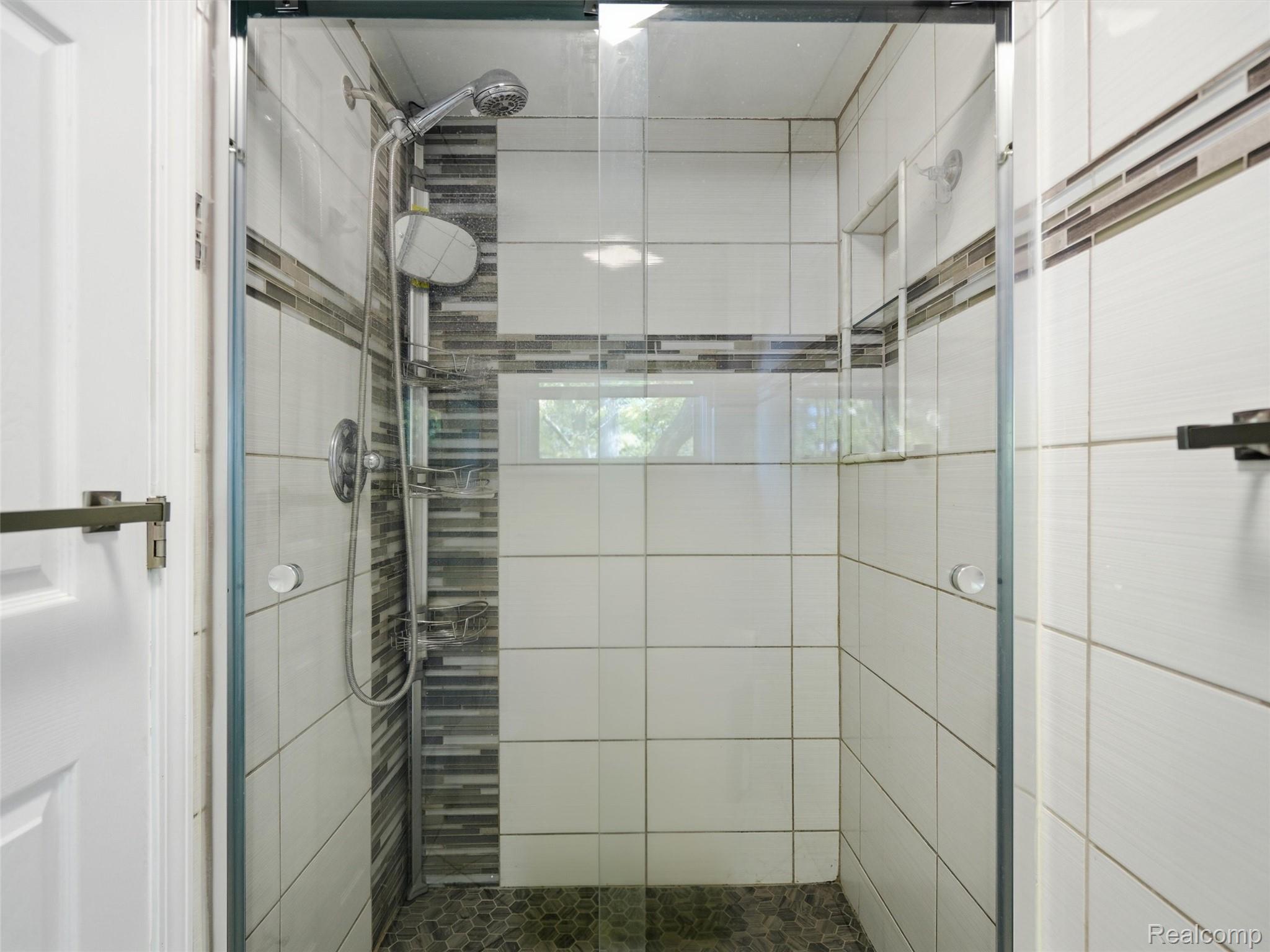 Walk-in shower with clear glass doors, large white tiles, and a handheld showerhead.