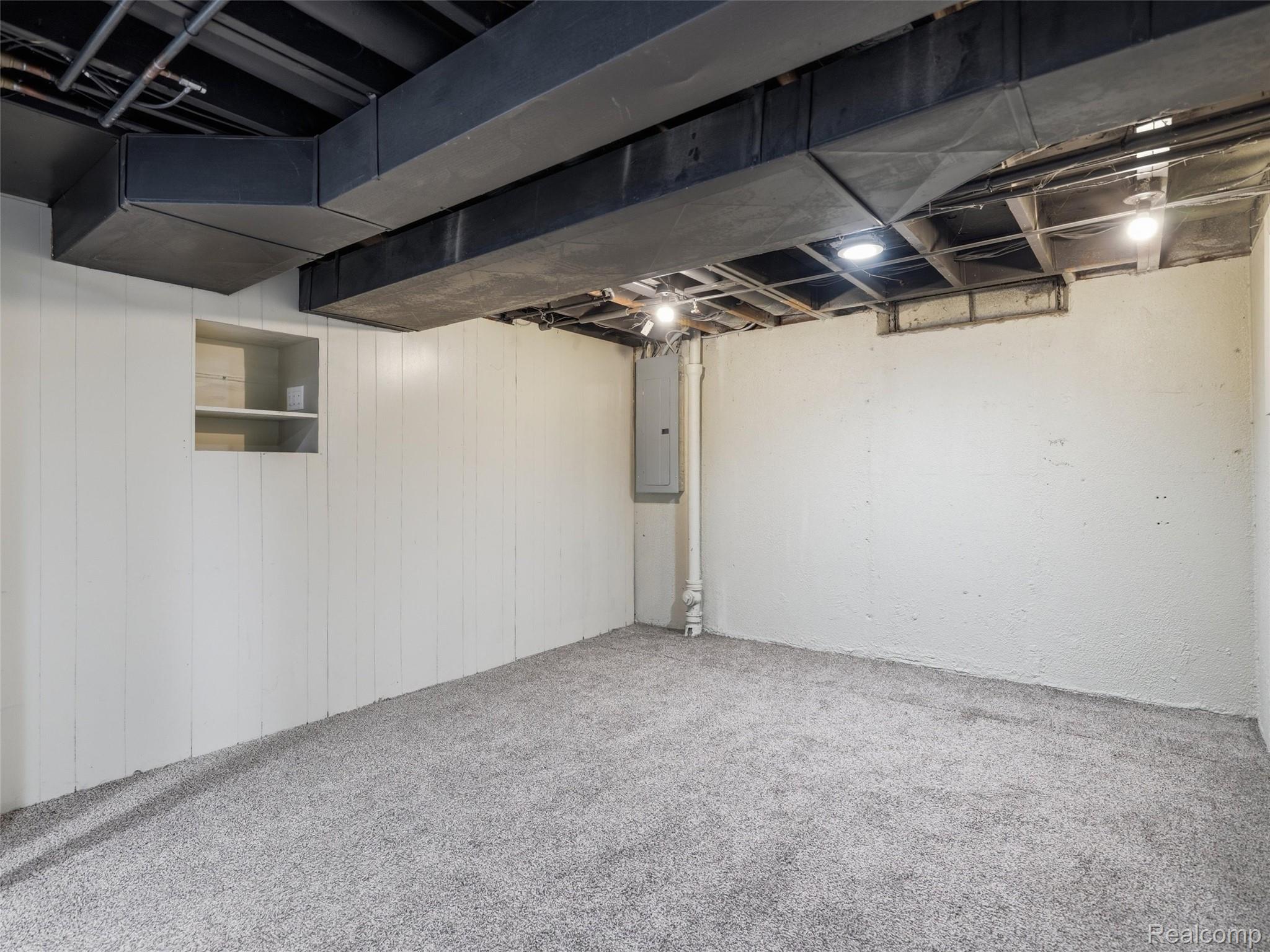 Unfinished basement room with gray carpet, exposed ceiling ductwork, and a utility panel.