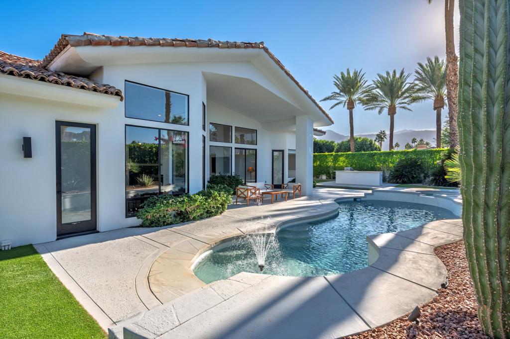 Modern white home beside a curved pool with a fountain, palm trees, and distant mountains.