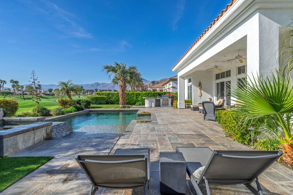 Backyard pool and stone patio beside a white stucco home, with a golf course and mountains beyond.