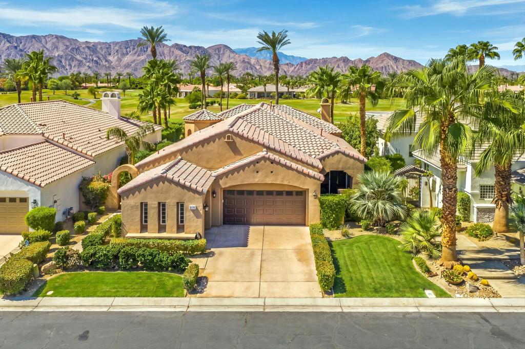 Tan stucco home with a tiled roof and two-car garage, backed by palms, golf-course grounds, and mountains.