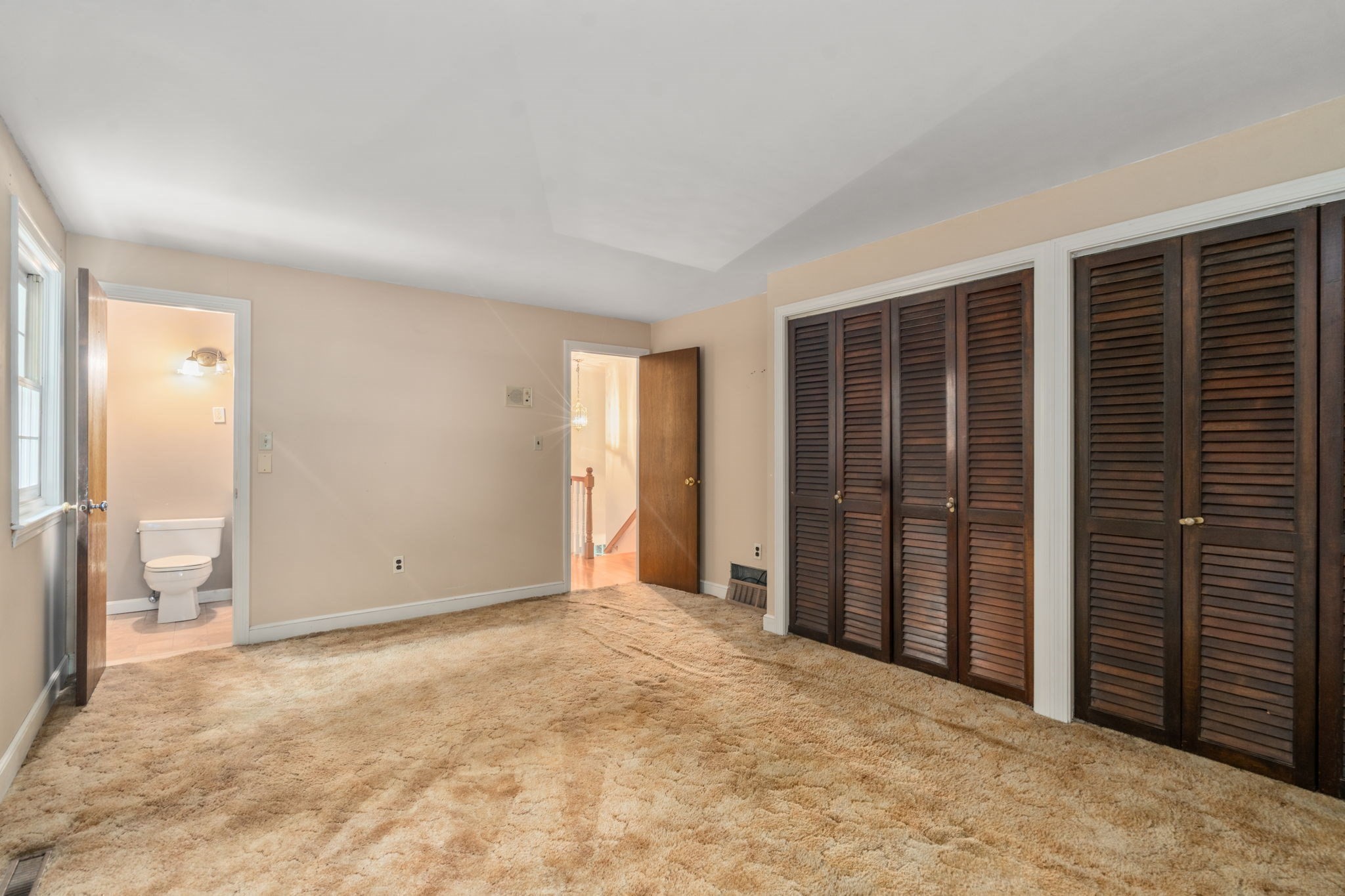 Empty bedroom with tan carpeting, dark louvered closets, and an adjoining half bathroom.