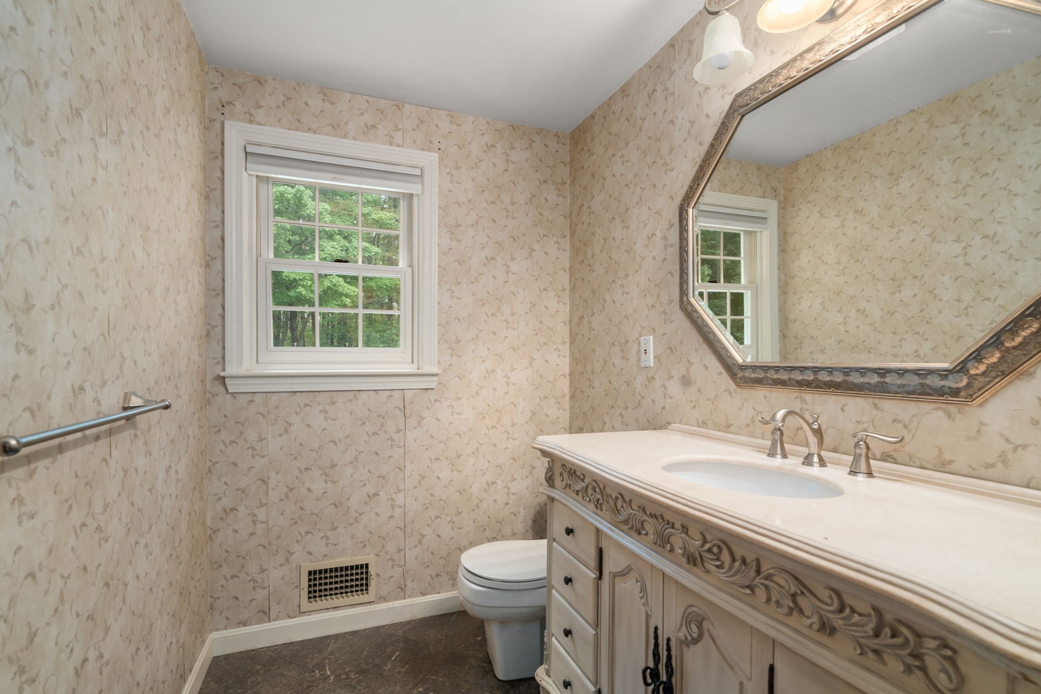 Bathroom with patterned beige walls, ornate vanity and mirror, and a window overlooking trees.