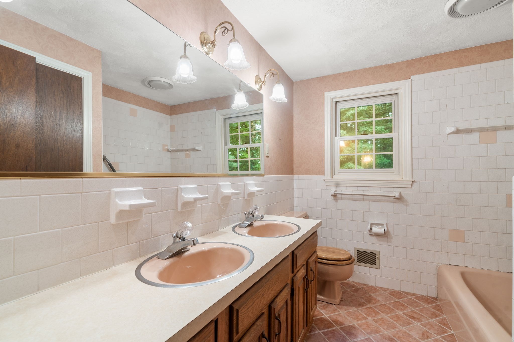 Bathroom with a long wood double vanity, peach sinks, white tile, and a window beside the tub.