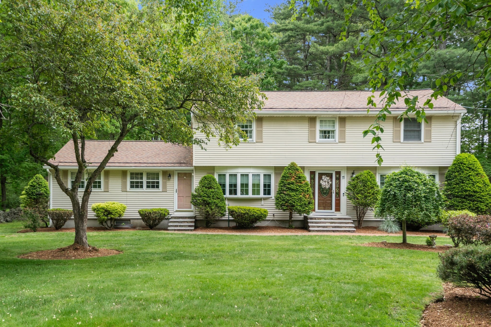 Beige split-level home with a broad green lawn, mature trees, and neatly trimmed shrubs.