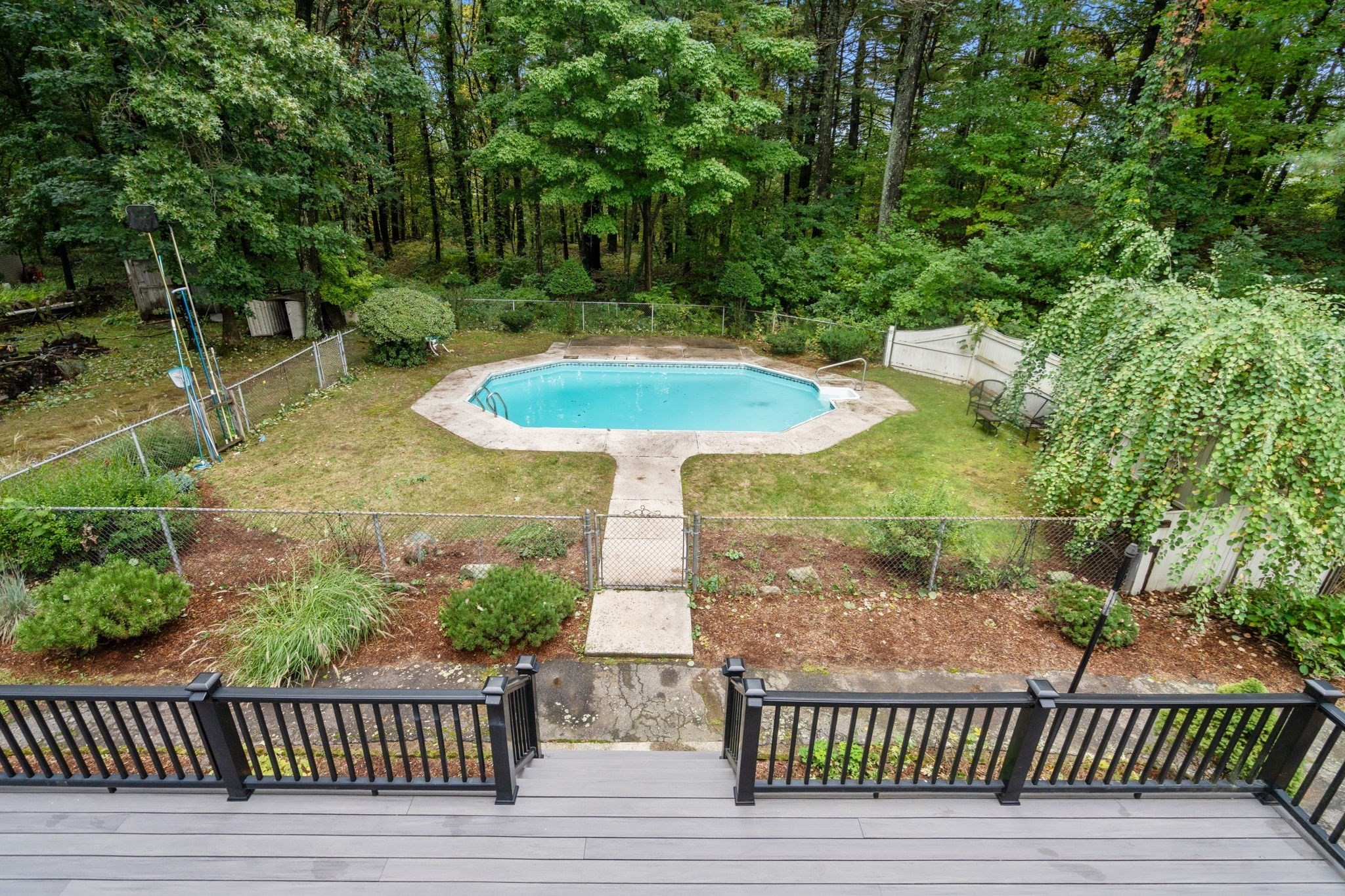 Backyard swimming pool viewed from a deck, with a fenced patio and wooded surroundings.
