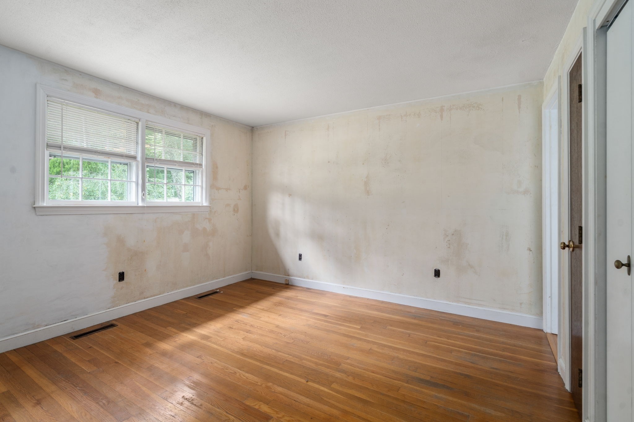 Vacant room with patchy walls, hardwood floors, and two windows with blinds.