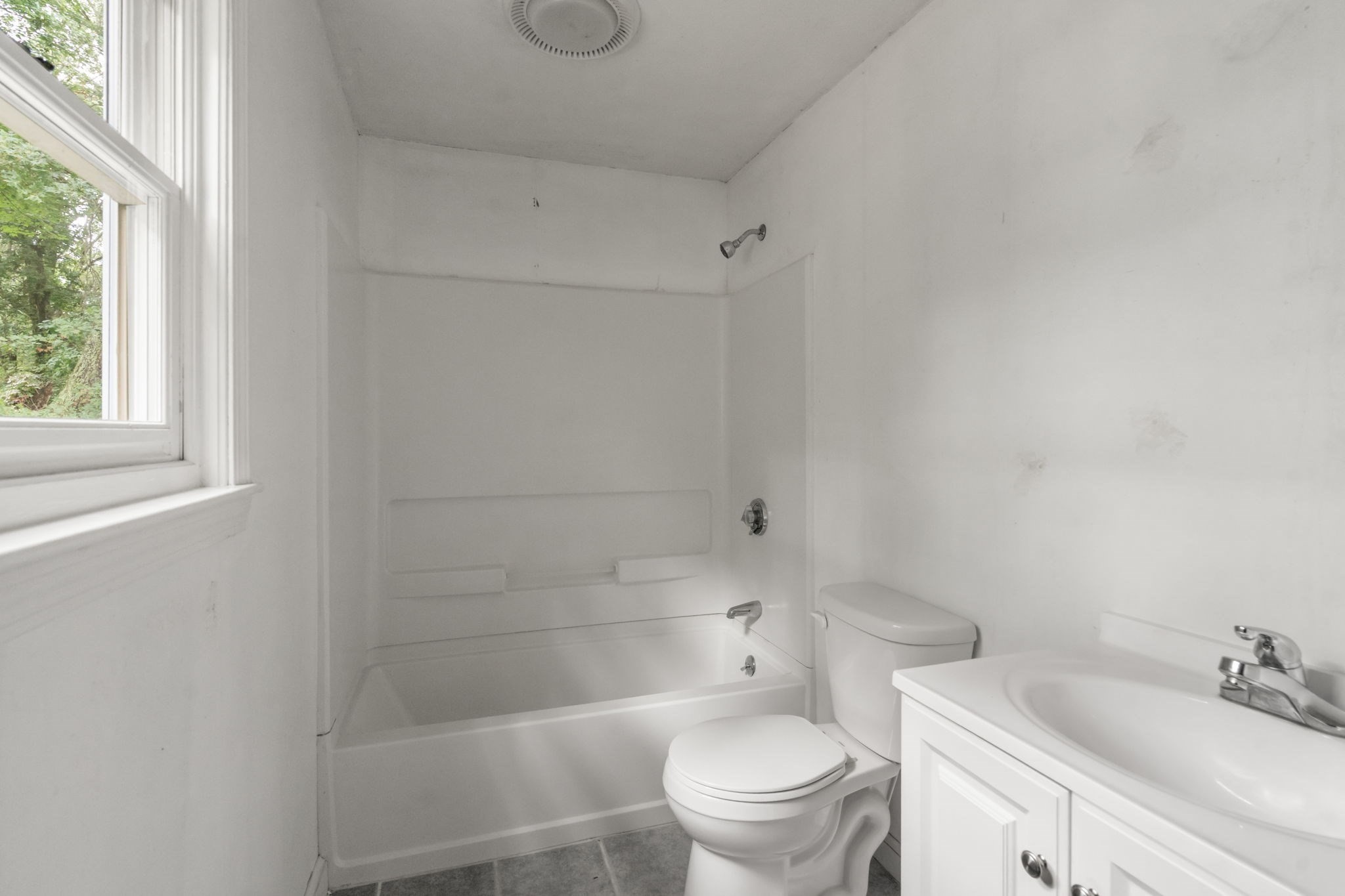 Bathroom with a white tub and shower, toilet, and vanity beside a window overlooking trees.