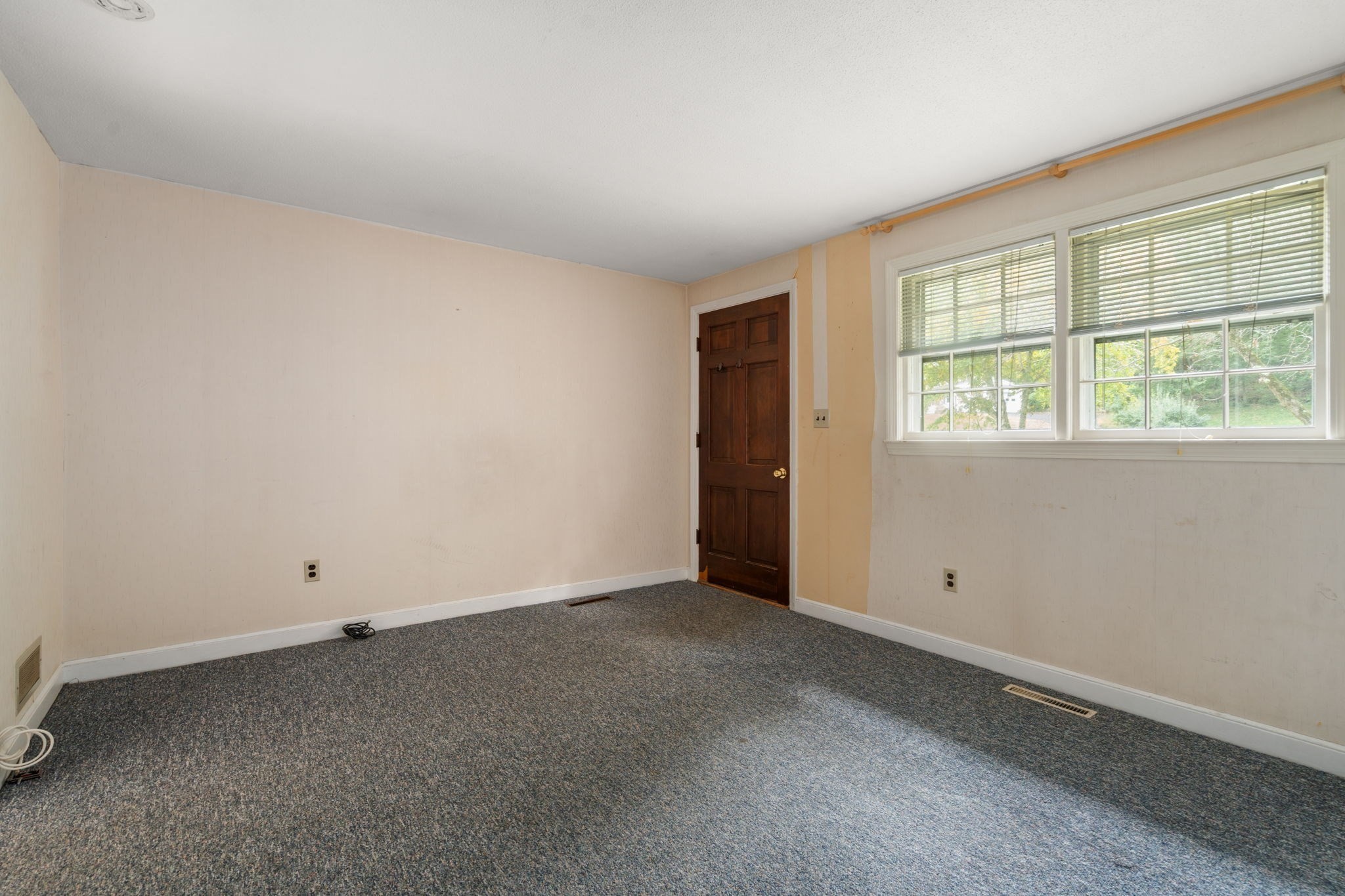 Empty carpeted room with beige walls, a dark wood door, and two wide windows with blinds.