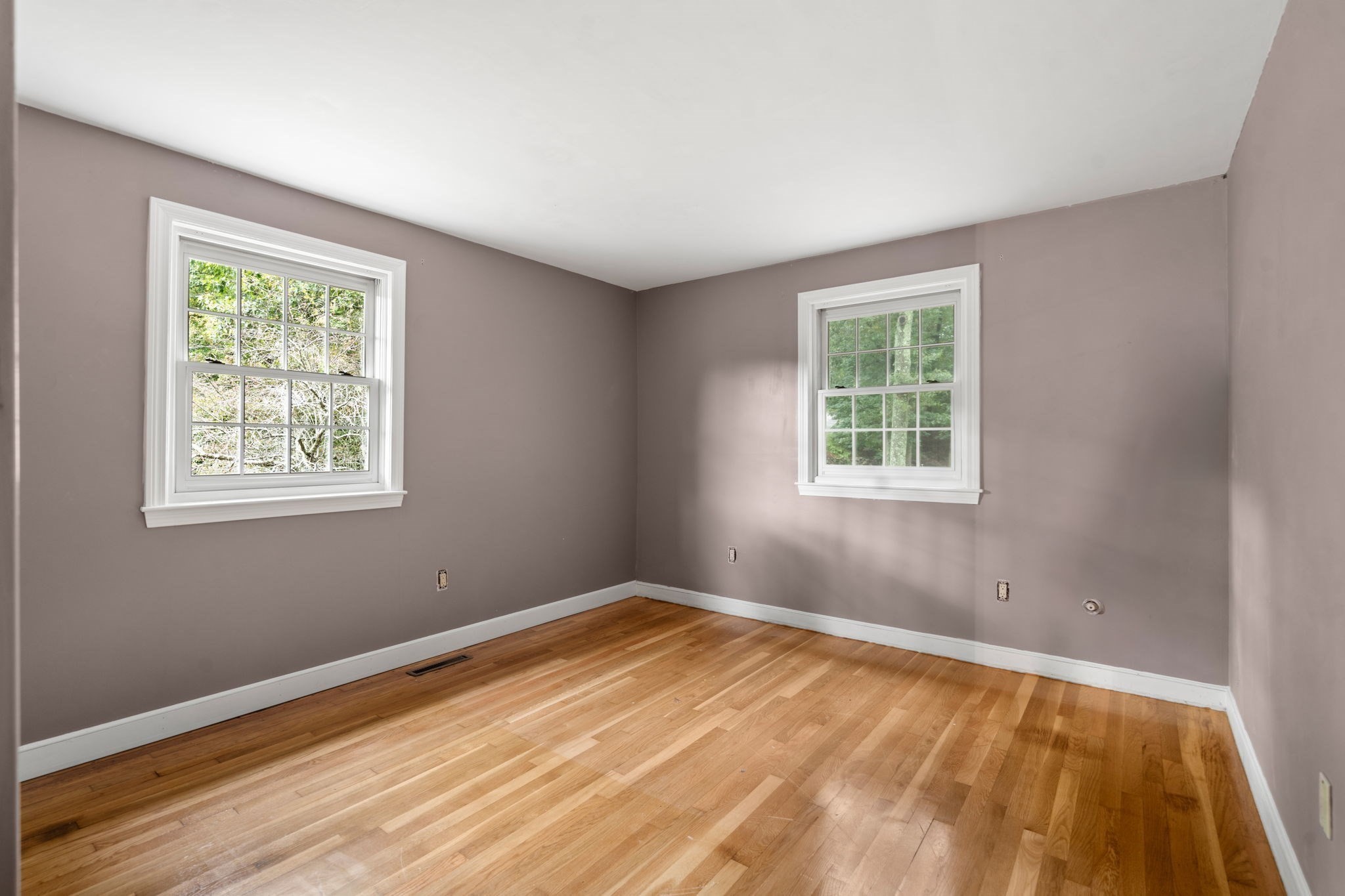 Empty room with polished hardwood floors, taupe walls, and two white-trimmed windows.