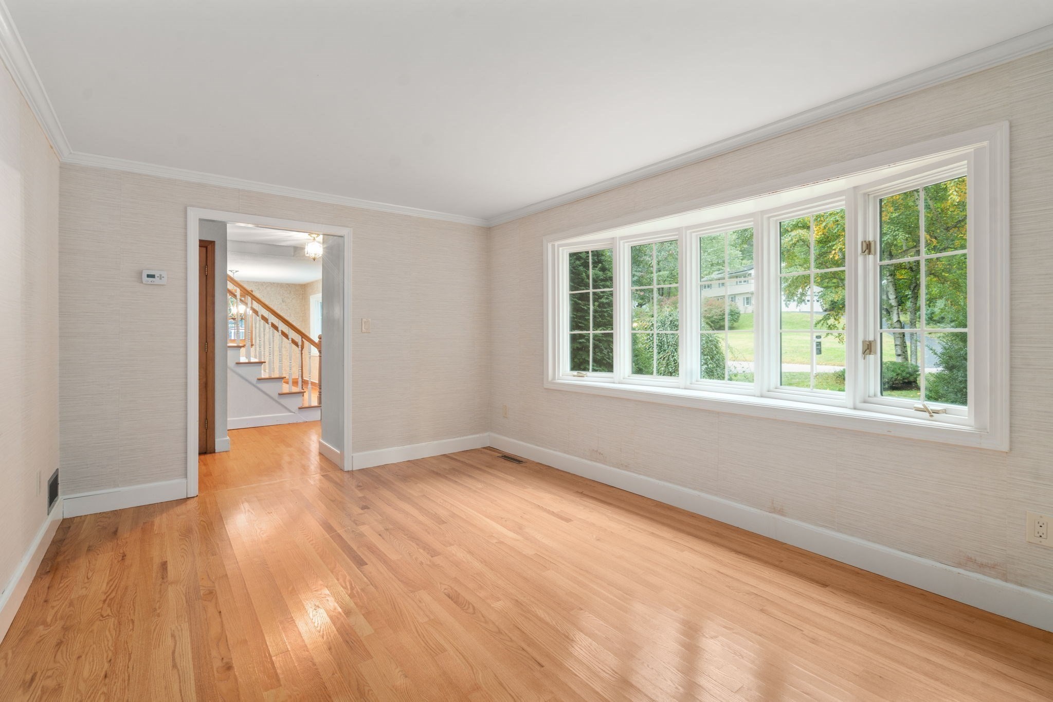 Empty room with hardwood floors and a wide row of windows overlooking a leafy yard.
