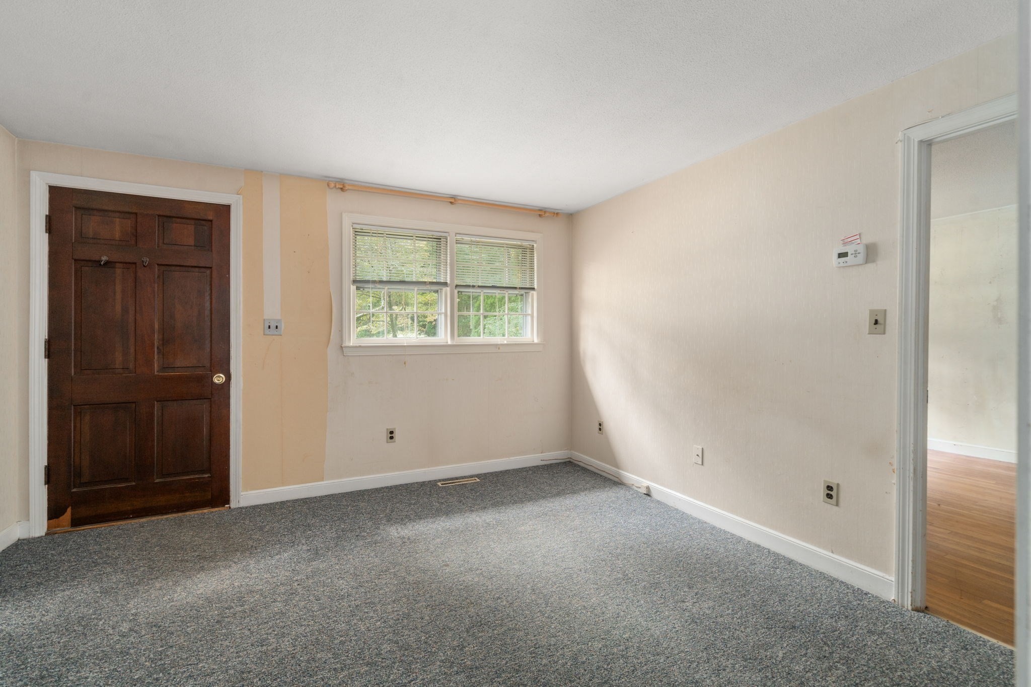 Empty carpeted room with a dark wood door, paired windows, and an adjoining hardwood-floored room.