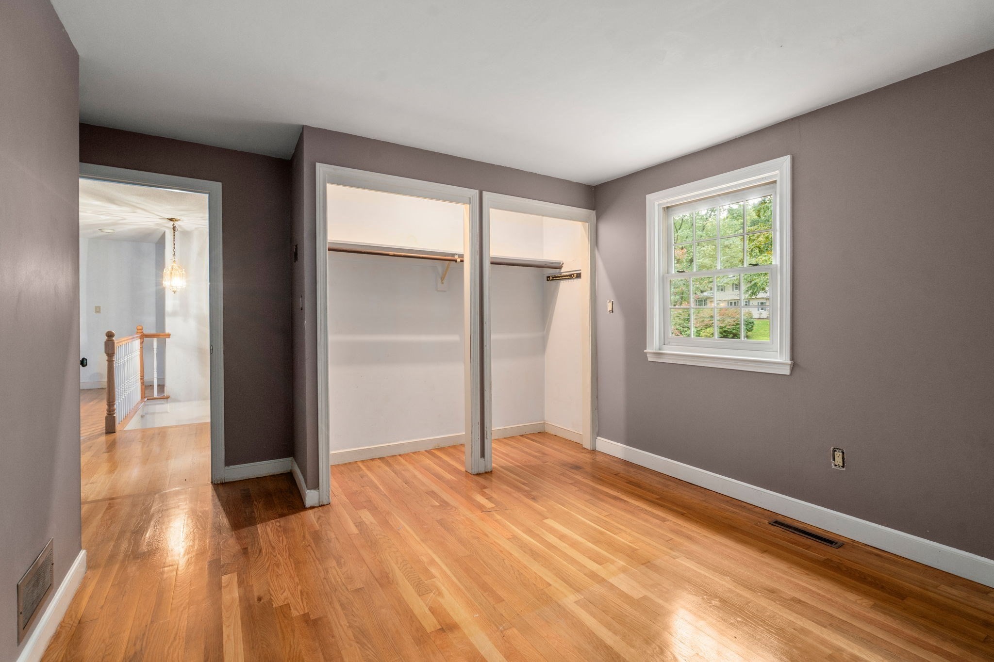 Empty bedroom with hardwood floors, two open closets, and a window overlooking trees.