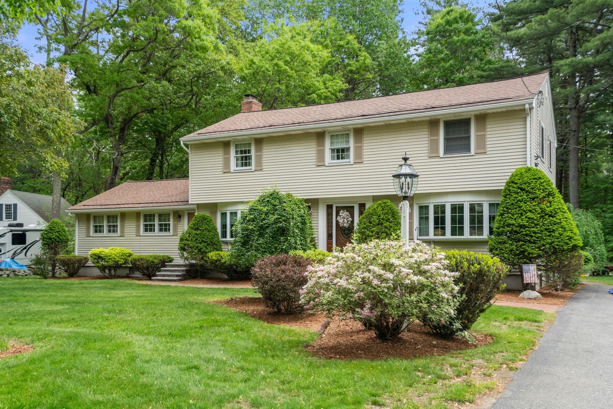 Beige two-story home with flowering shrubs, mature trees, and a landscaped front lawn.