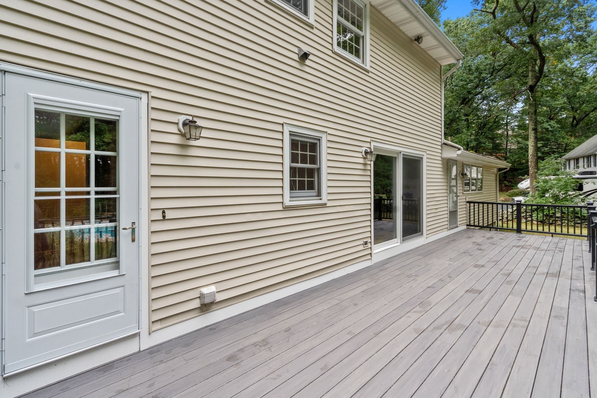 Spacious gray deck beside a beige-sided house, with black railing and mature trees beyond.