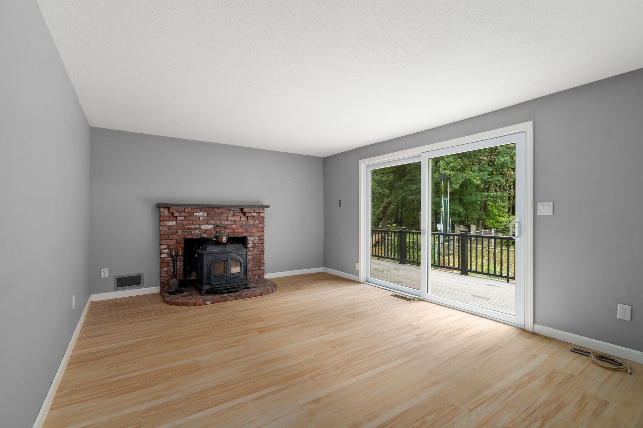 Empty room with a brick fireplace and wood stove, light wood floors, and sliding doors to a wooded deck.
