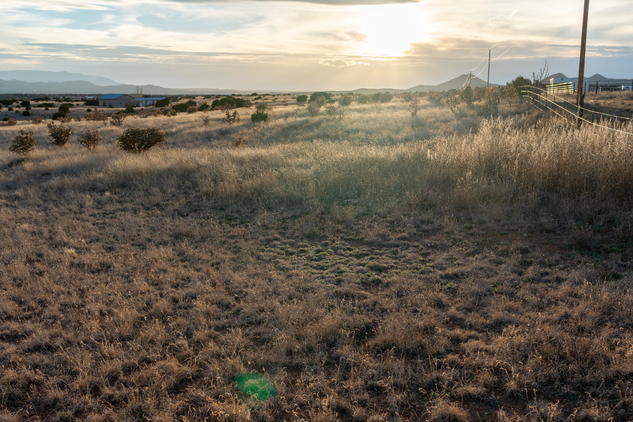 Vacant grassy parcel with scattered shrubs, a fence, and distant mountains at sunset.