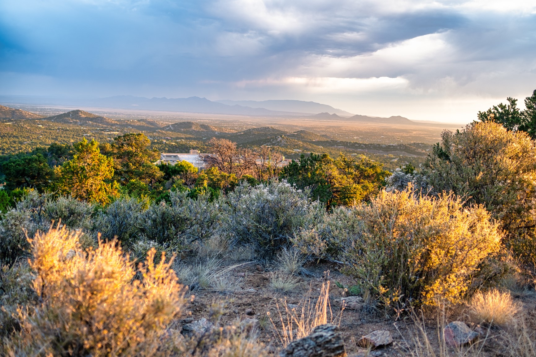 Mountain valley view from a rocky hillside, with a home partly visible among shrubs.